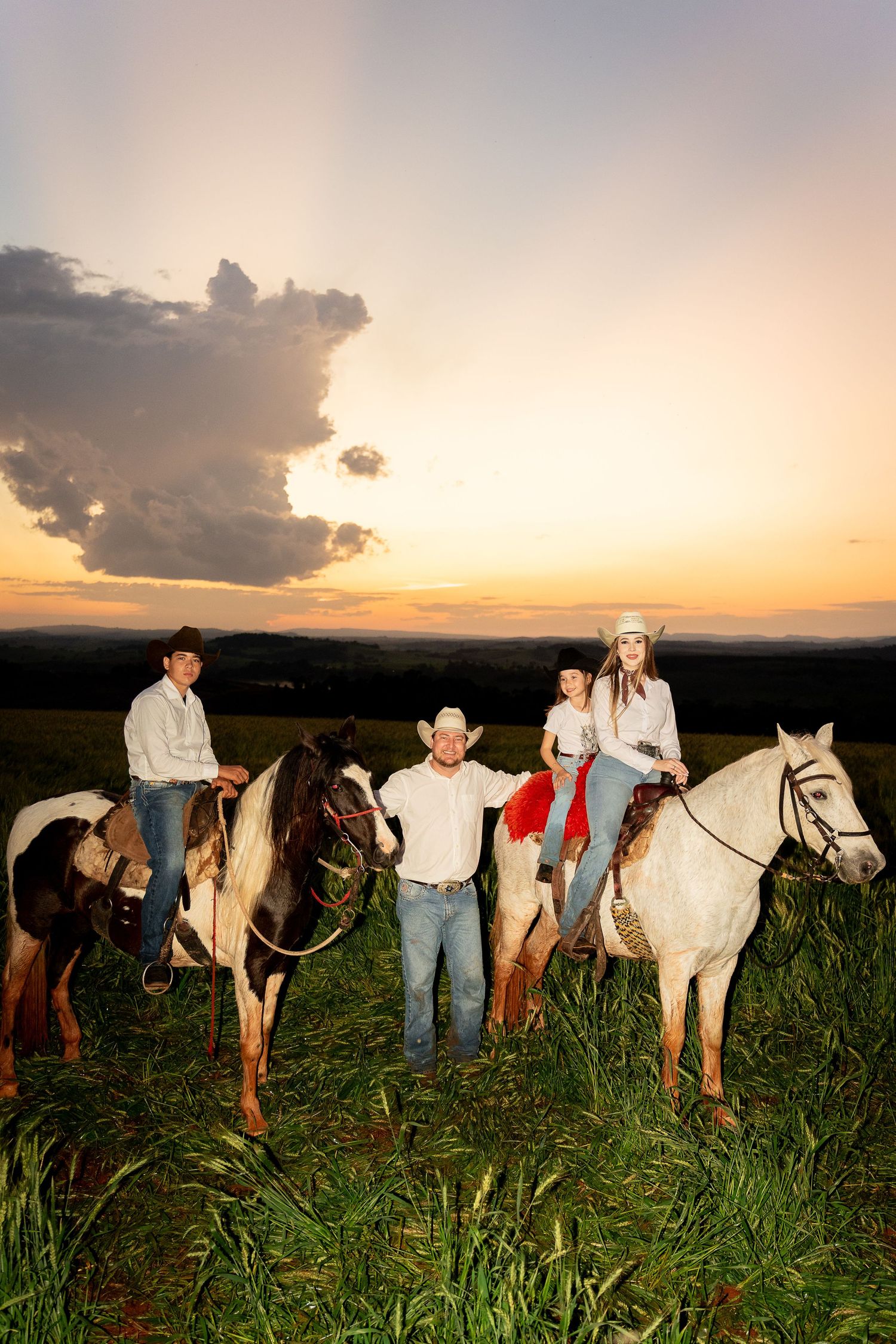 Family of four with horses in a green field at sunset, wearing western attire including cowboy hats.