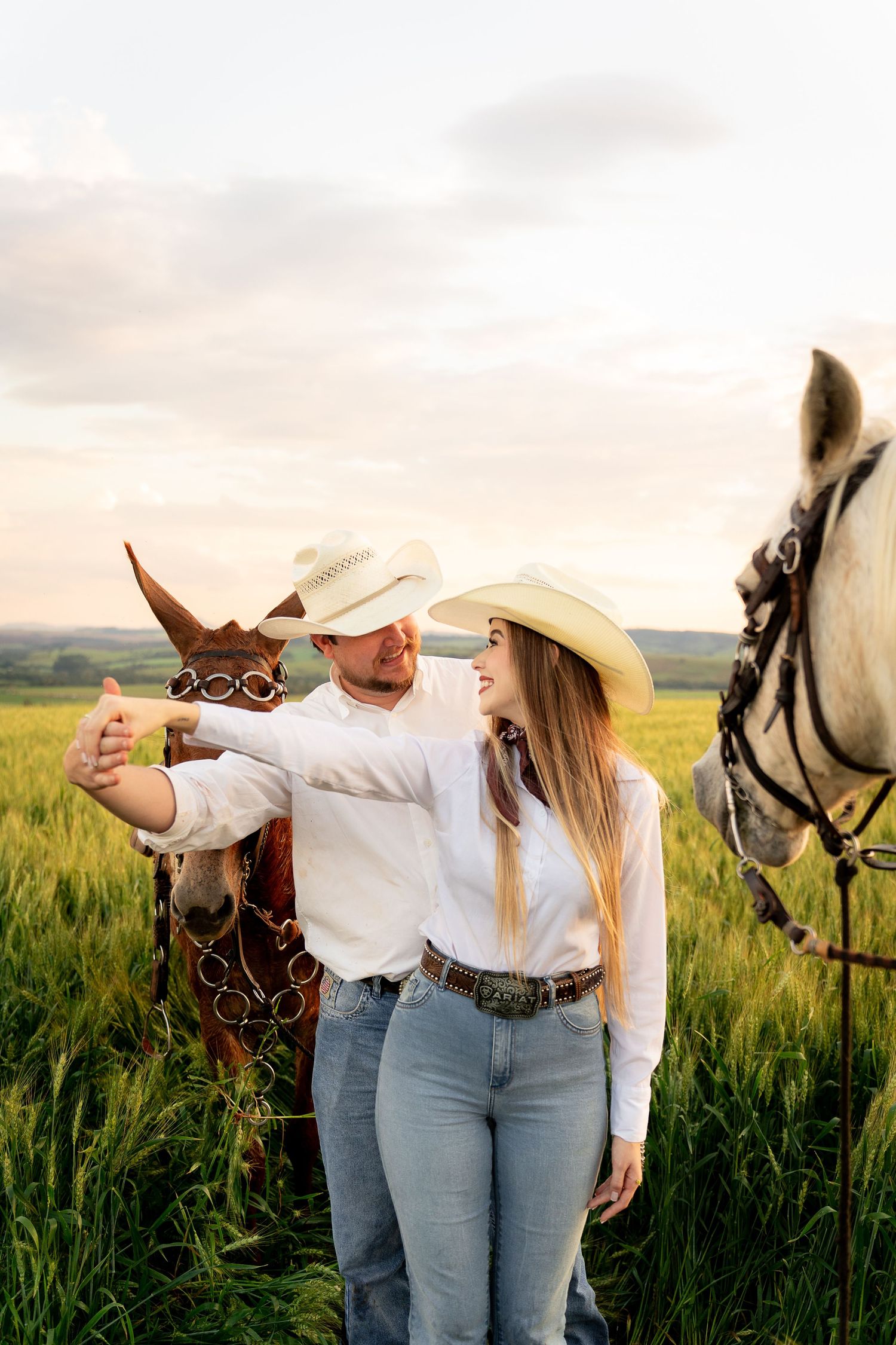 Couple in cowboy hats taking a selfie with brown and white horses in a green field at sunset.