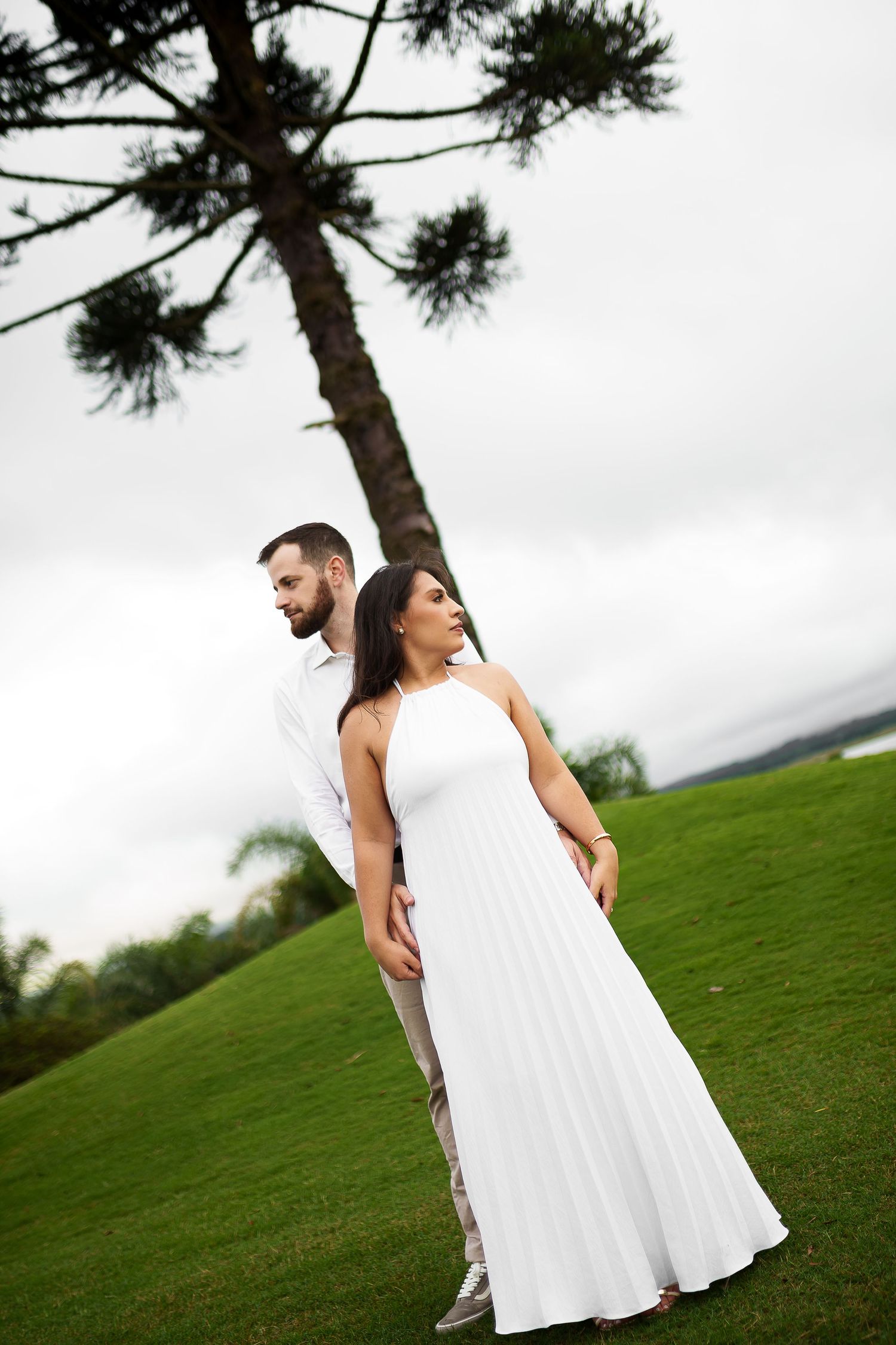 Couple in white attire standing on lush green lawn with tall pine tree backdrop on overcast day.