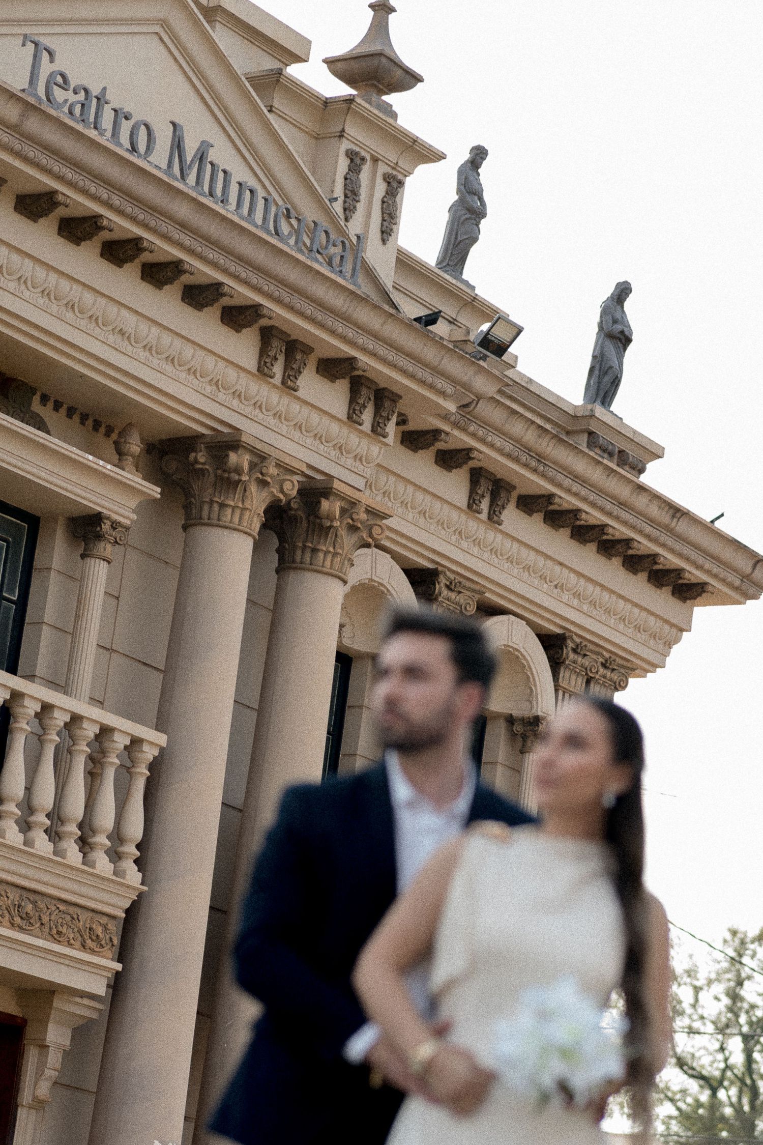 Bride and groom pose in soft focus before the ornate Teatro Municipal building with classical columns and statues.