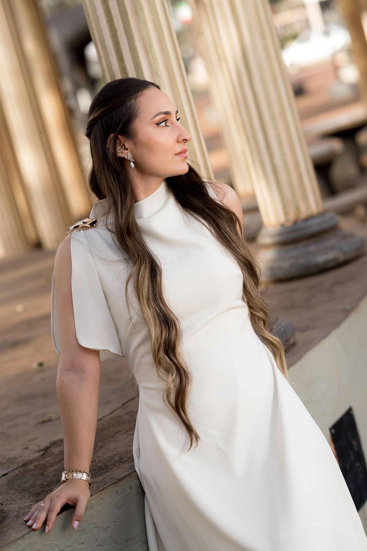 Woman in white gown with long wavy hair poses gracefully among classical columns in an elegant outdoor setting.