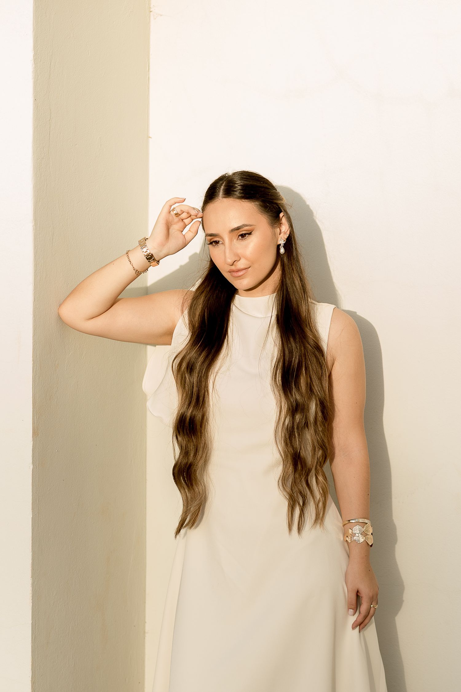 Woman in white sleeveless dress with long wavy hair poses gracefully against a cream wall wearing gold bracelets.