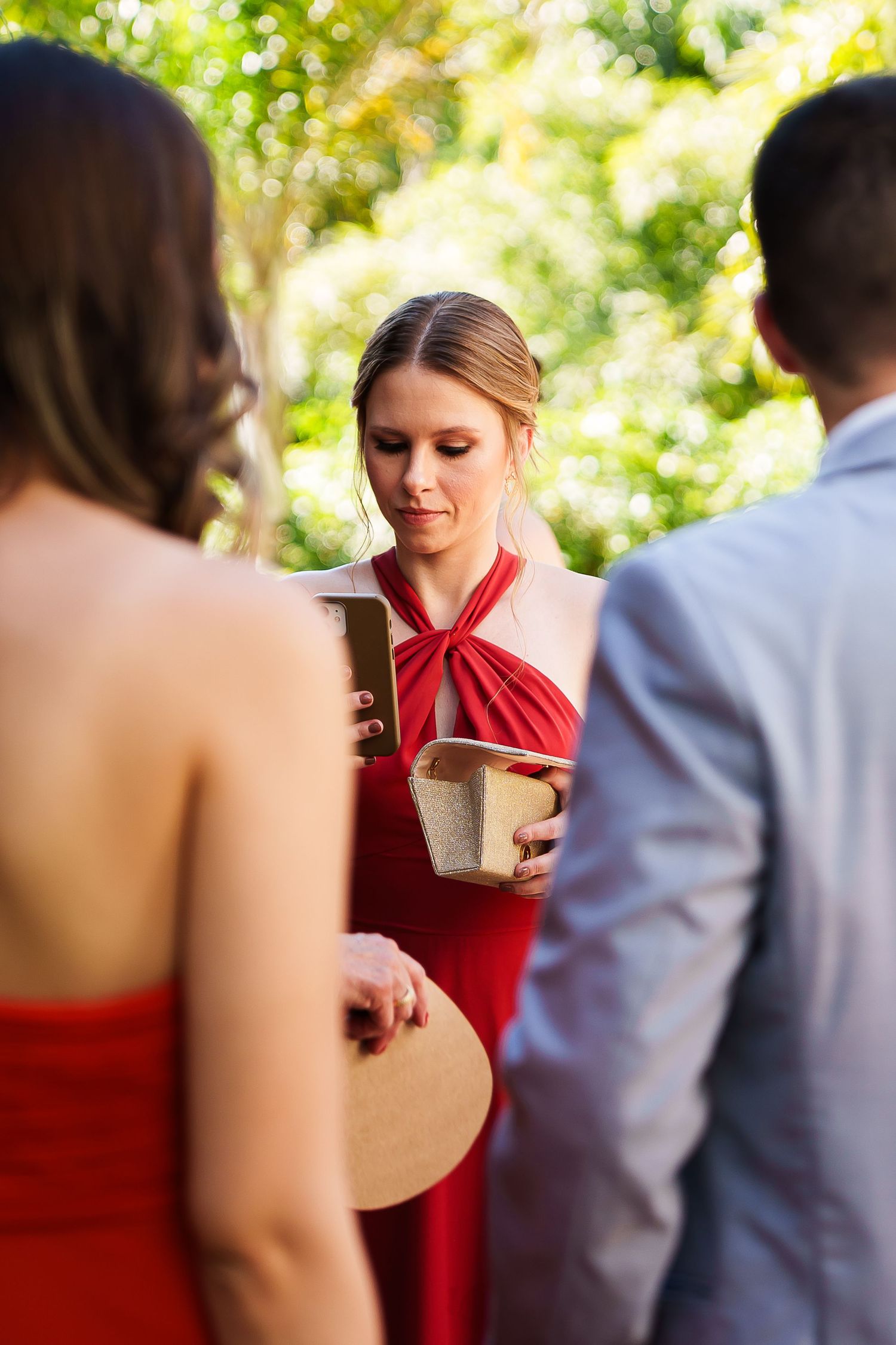 Woman in red dress holding boxes at an outdoor wedding ceremony with guests in foreground.