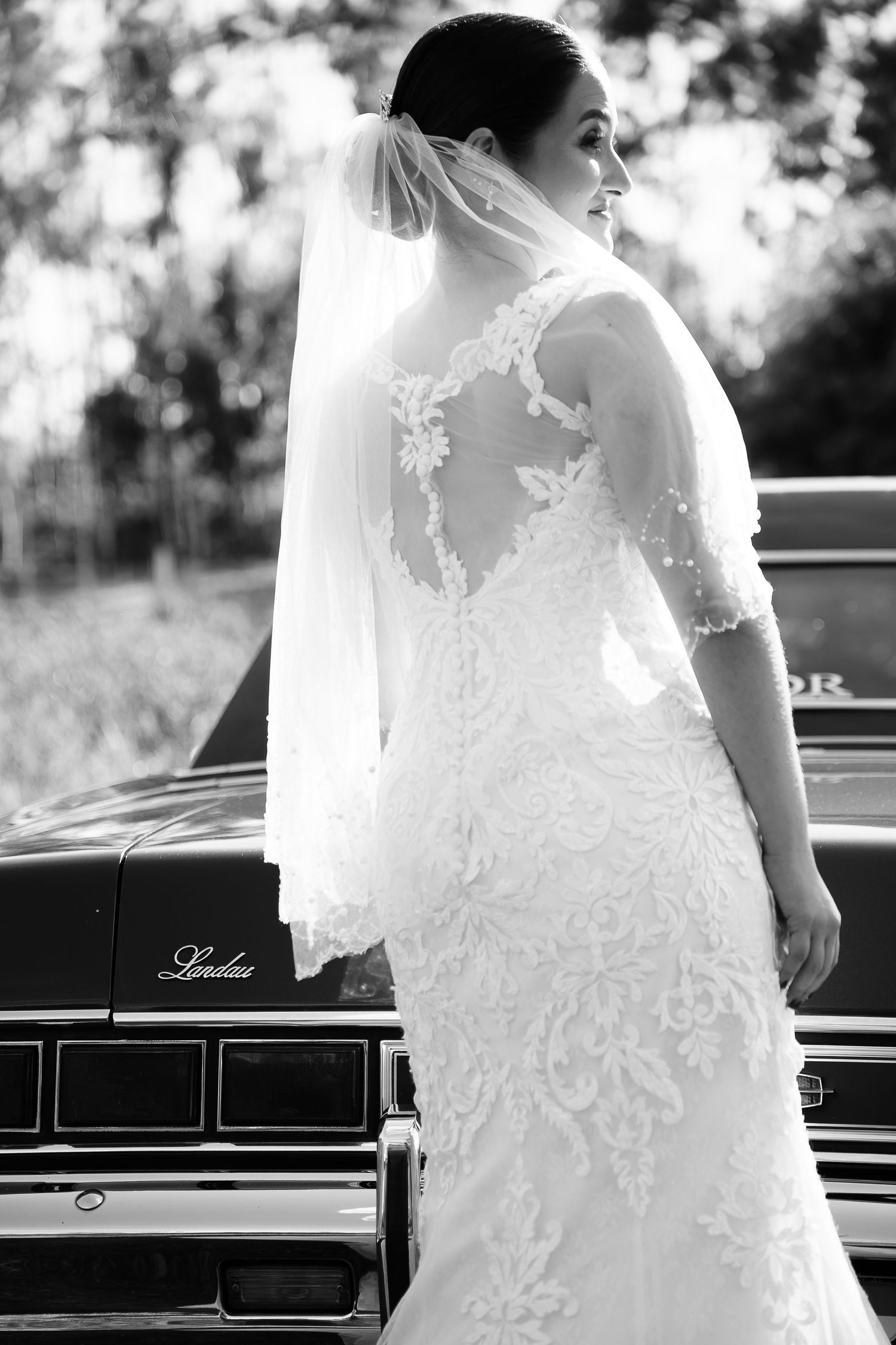 Bride in lace wedding gown with veil stands beside vintage Lincoln car in elegant black and white portrait.
