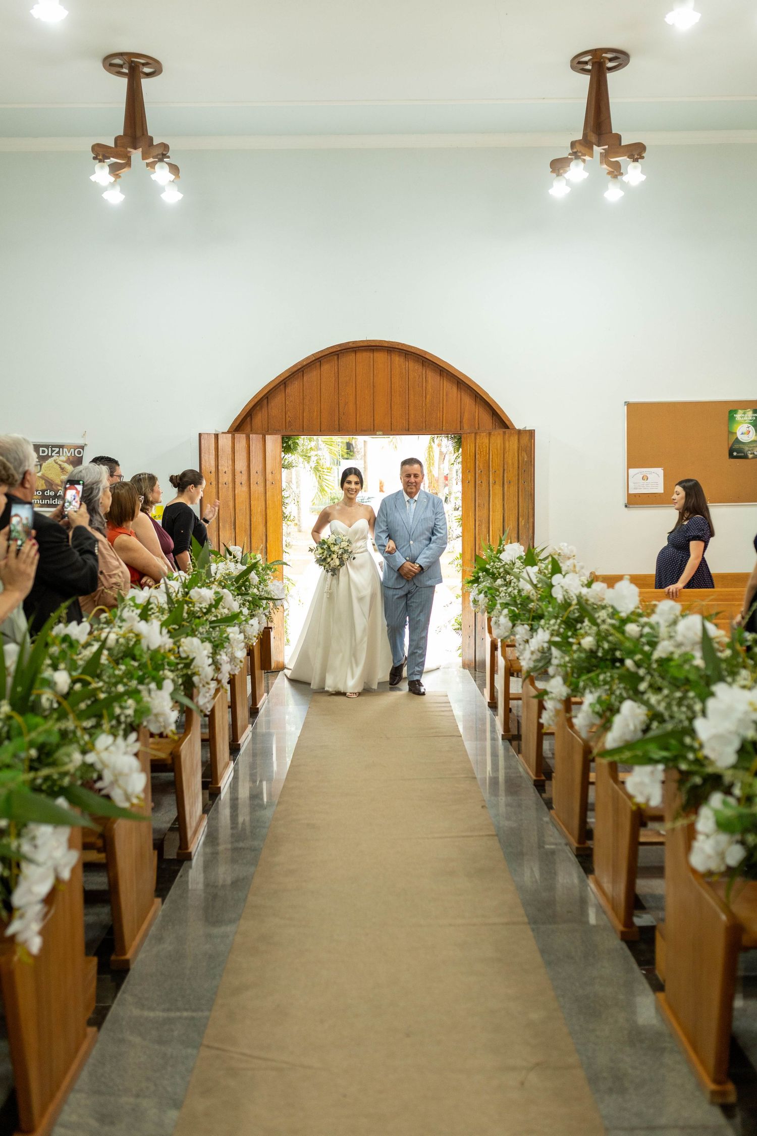 Bride walks down the aisle with escort through floral-lined church pews during elegant wedding ceremony.