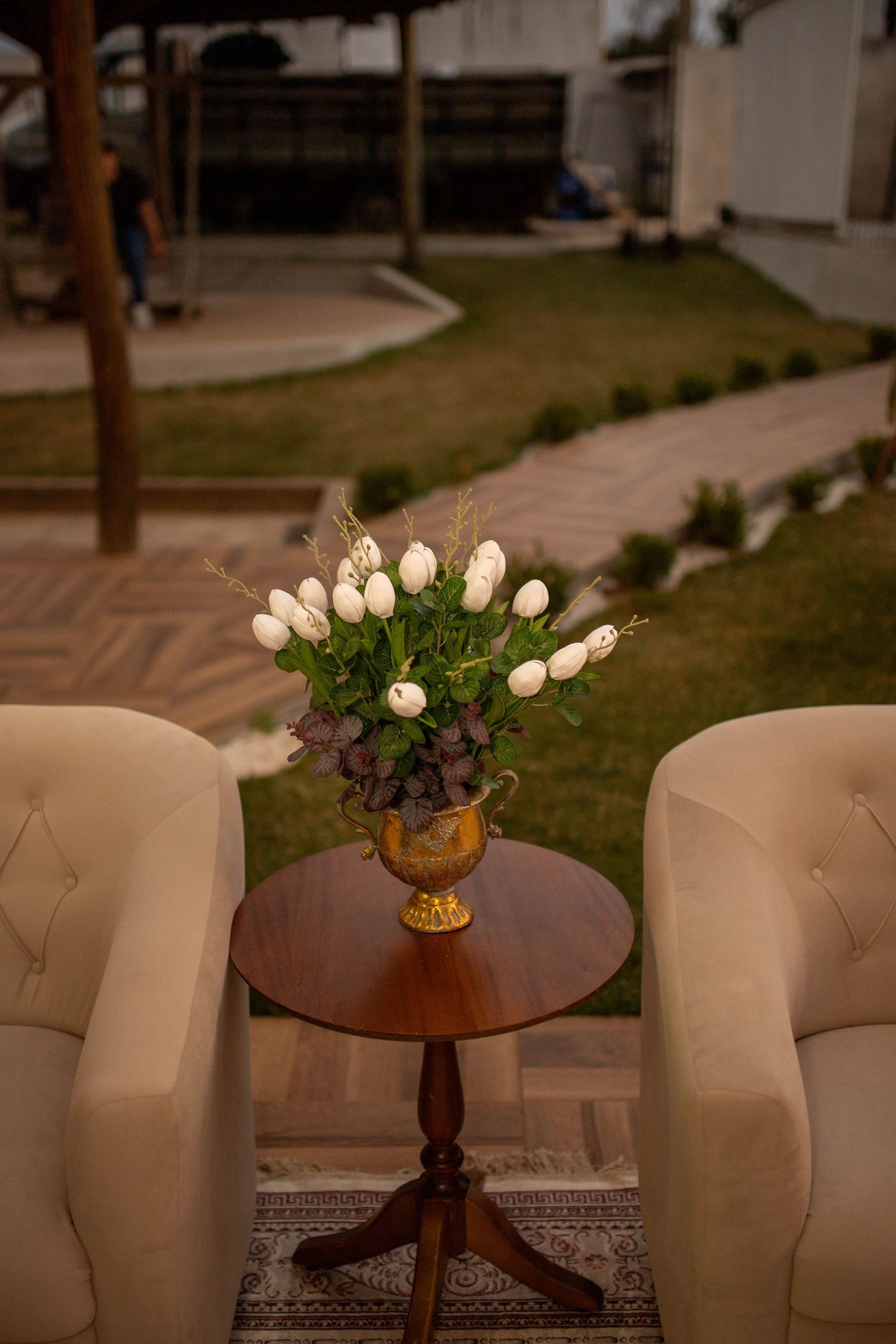 Elegant white tulips in a gold vase on a wooden pedestal table flanked by cream armchairs in a garden setting.