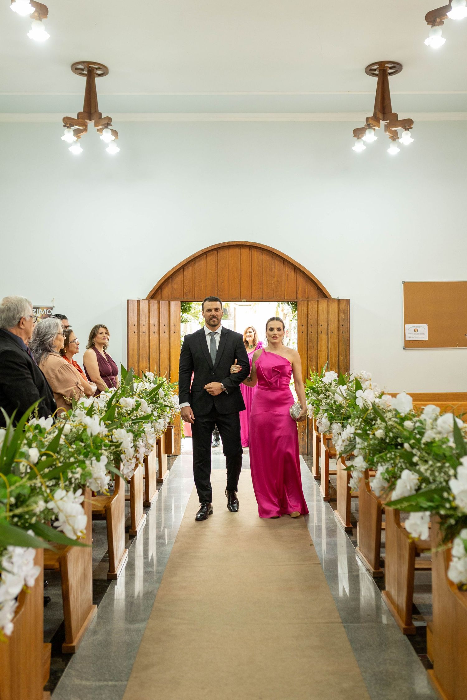 A couple walks down a flower-lined church aisle, the woman in a pink gown and man in a dark suit, during a wedding ceremony.