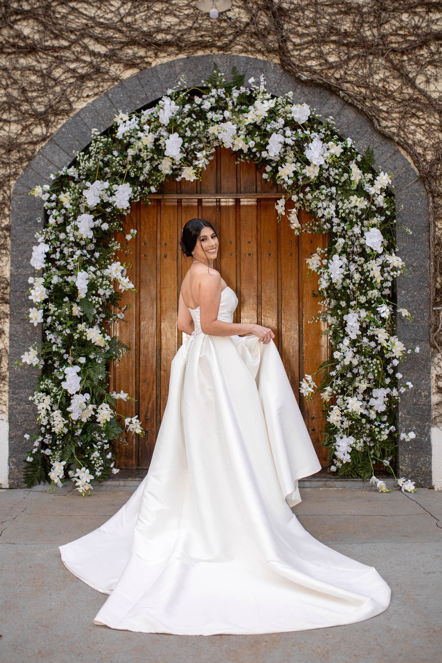 Bride in elegant white ball gown with train poses under floral arch of white roses at wooden door.