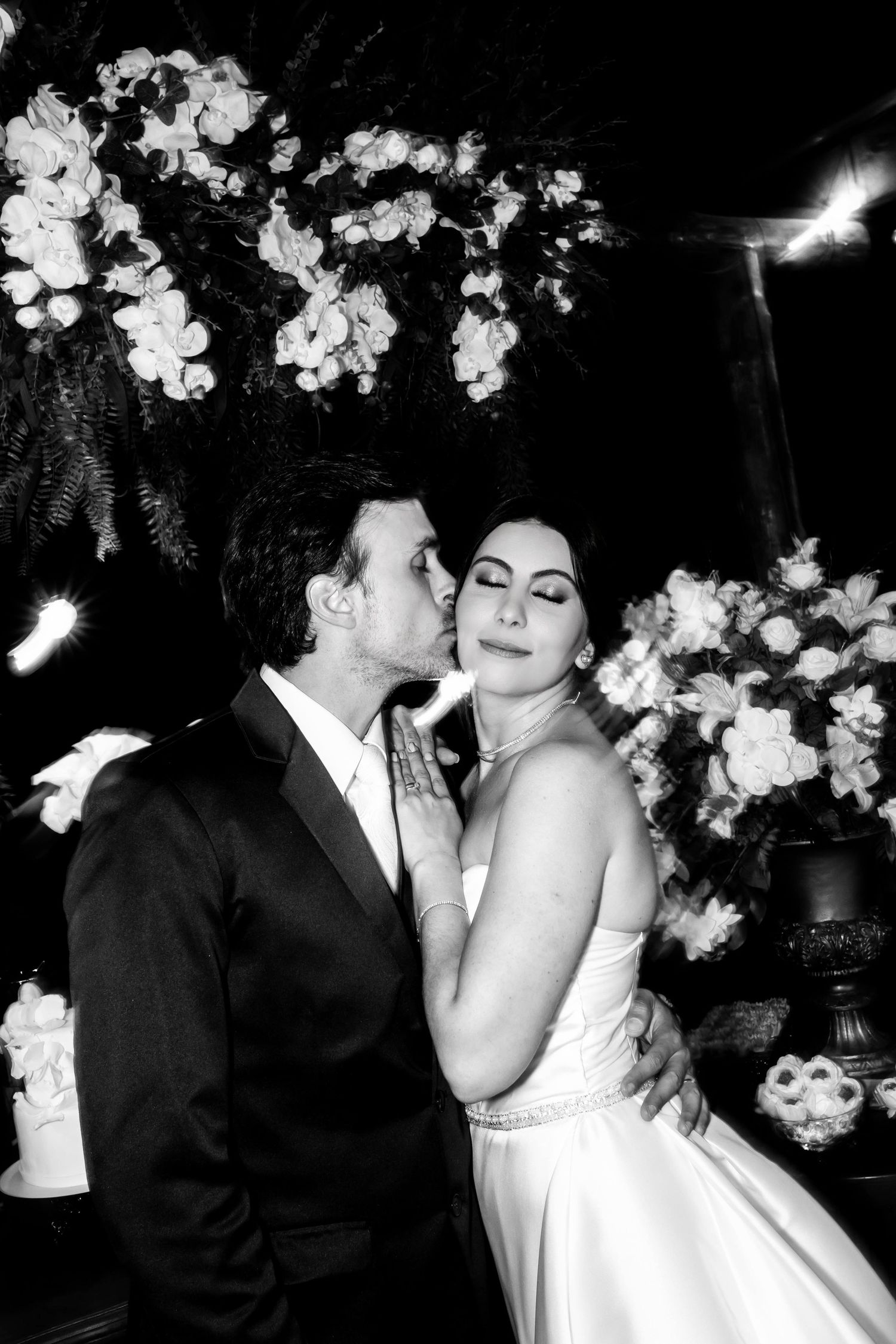 Black and white wedding photo of groom kissing bride on cheek surrounded by white floral arrangements.