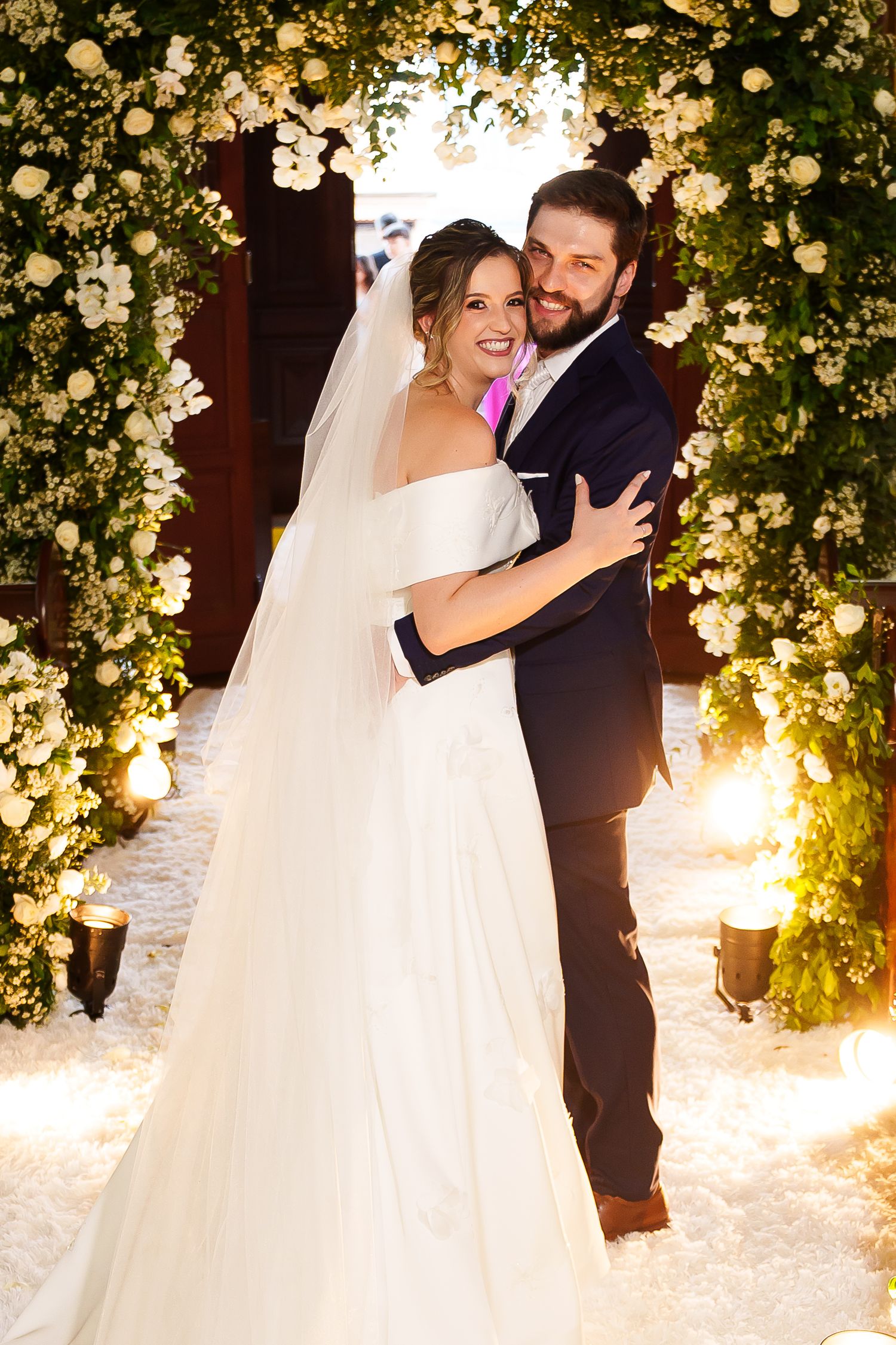 A smiling bride and groom embrace under a floral arch adorned with white flowers and greenery at their wedding ceremony.