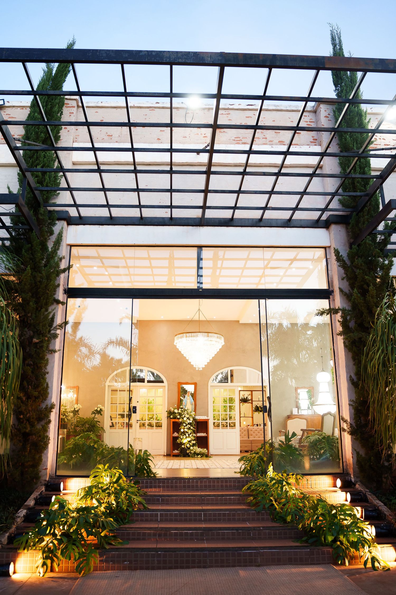 Elegant venue entrance with glass walls, chandelier, brick steps, lush greenery, and a pergola overhead at dusk.