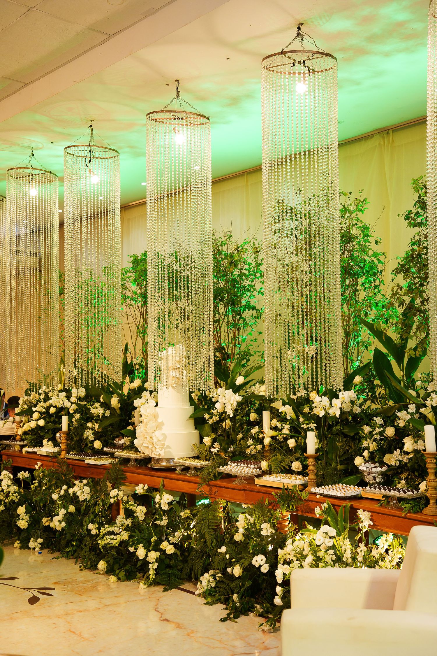 Elegant wedding reception with crystal chandelier curtains, white floral arrangements, and a tiered cake on a decorated table.