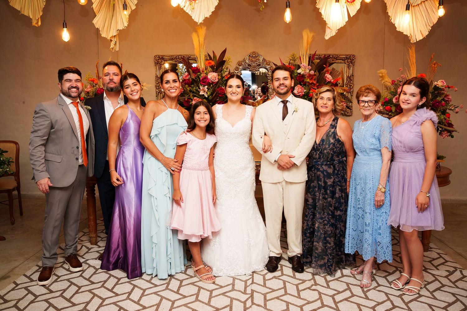 Wedding party poses together, bride in white gown, groom in tan suit, surrounded by family in colorful formal attire.