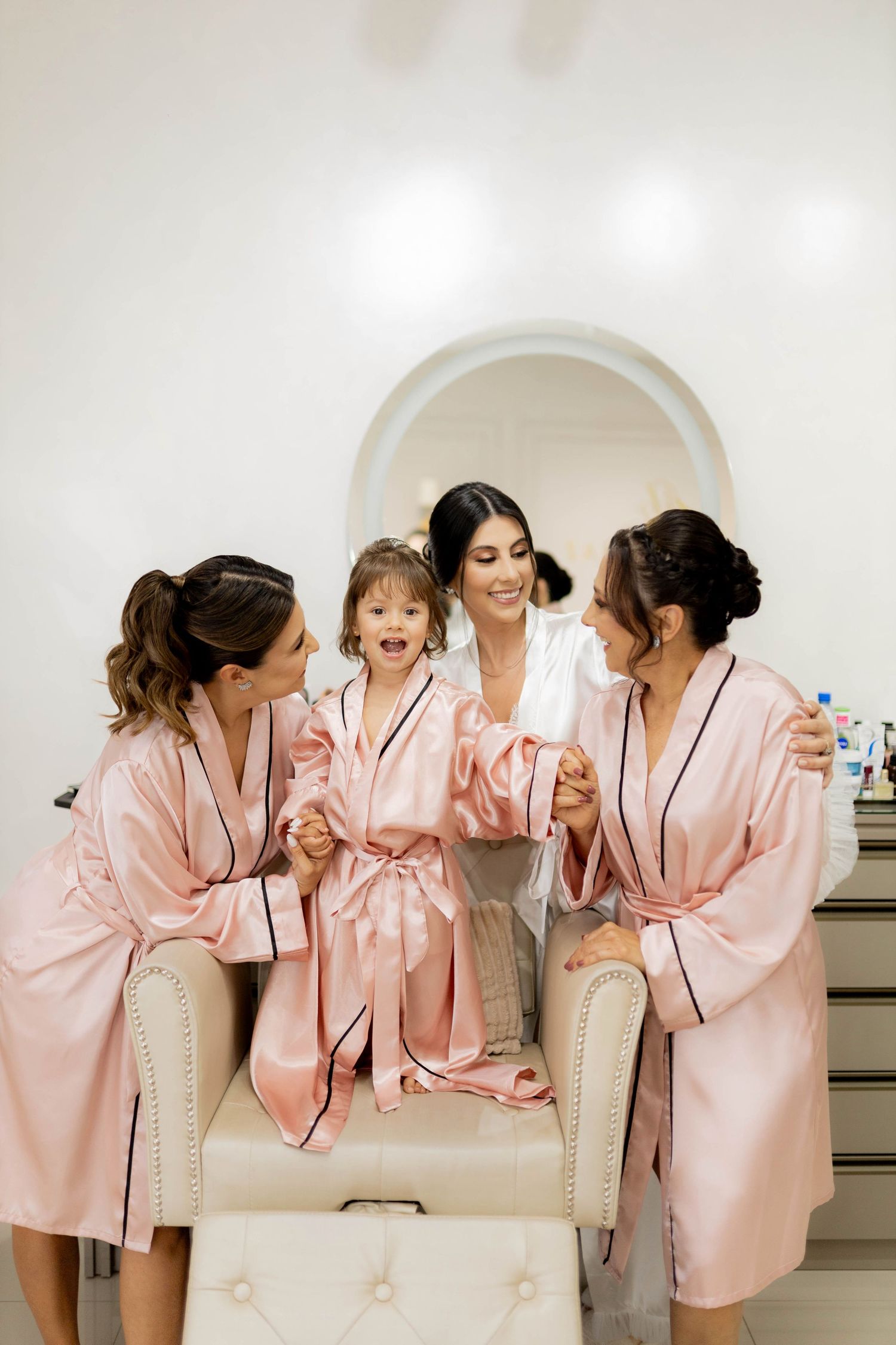 Bride and bridesmaids in pink satin robes laughing together with a flower girl during wedding morning preparations.