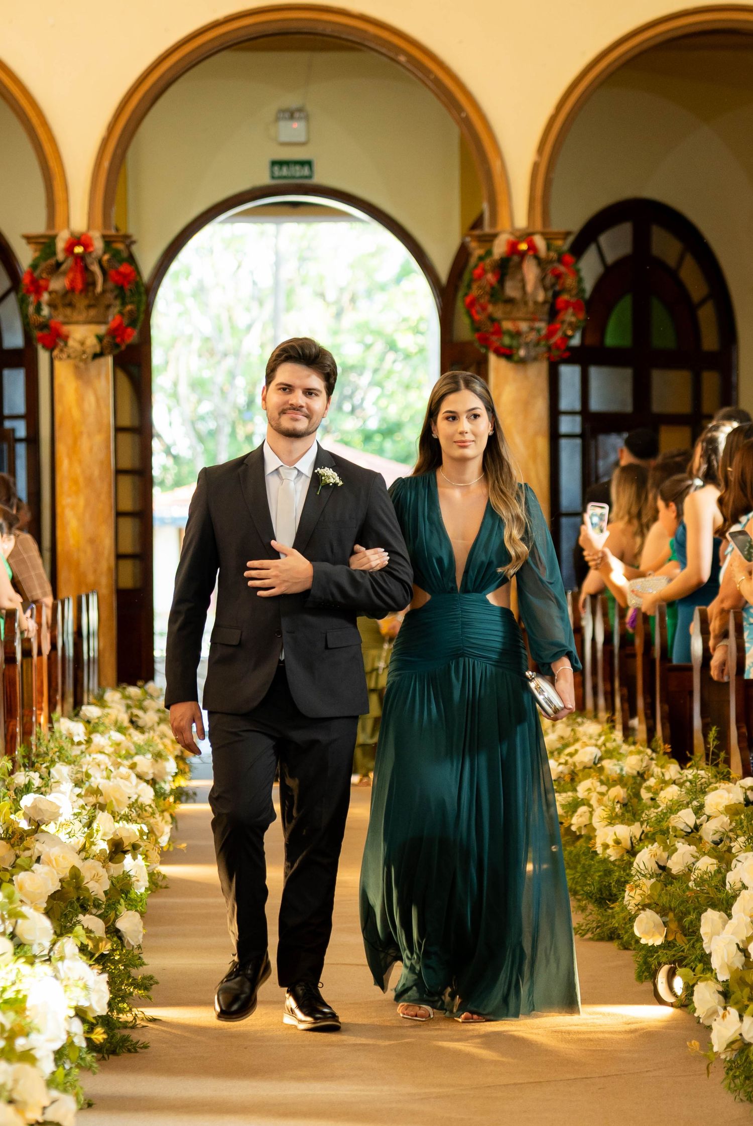 A couple walks down a flower-lined church aisle, man in black suit, woman in emerald green gown, at a elegant wedding ceremony.