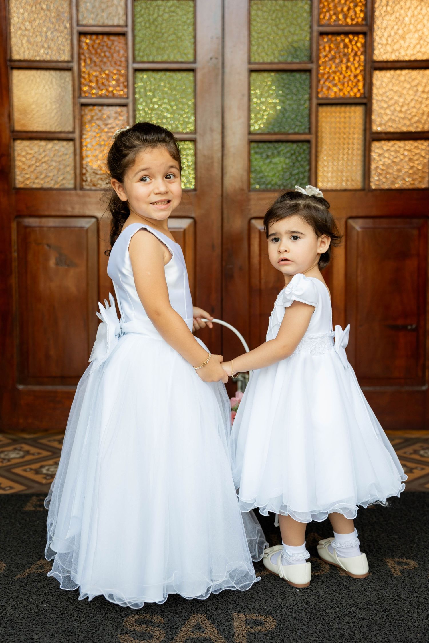 Two young flower girls in white dresses holding a basket near ornate wooden doors.