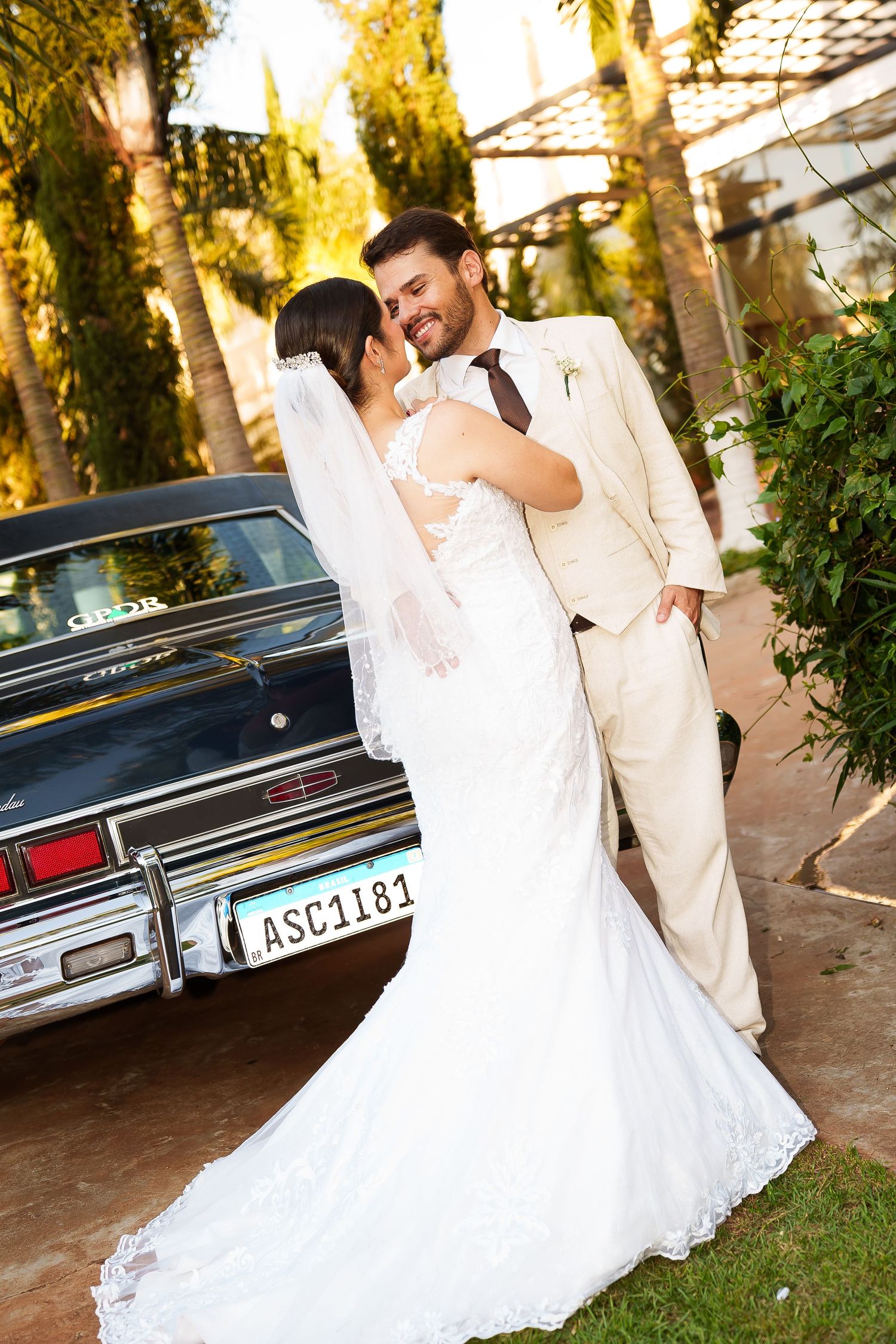 A bride in a white lace gown and groom in a cream suit share a romantic moment beside a classic dark blue car.