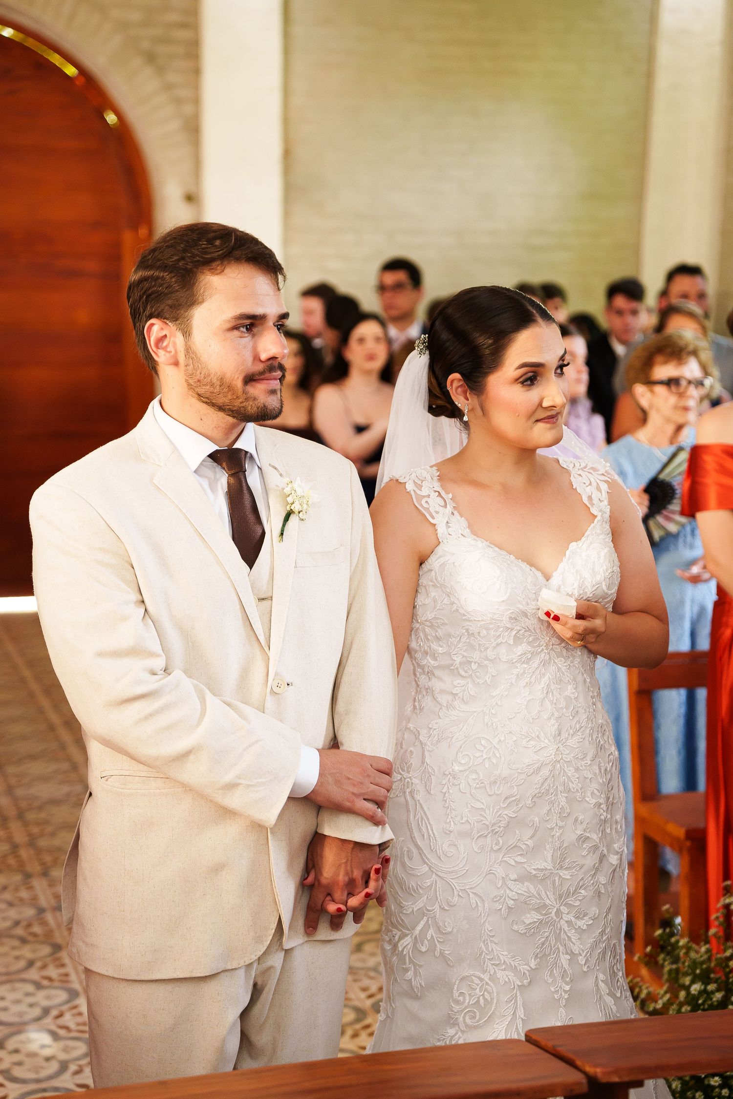 Bride and groom holding hands at wedding ceremony altar, groom in cream suit, bride in lace dress with veil.