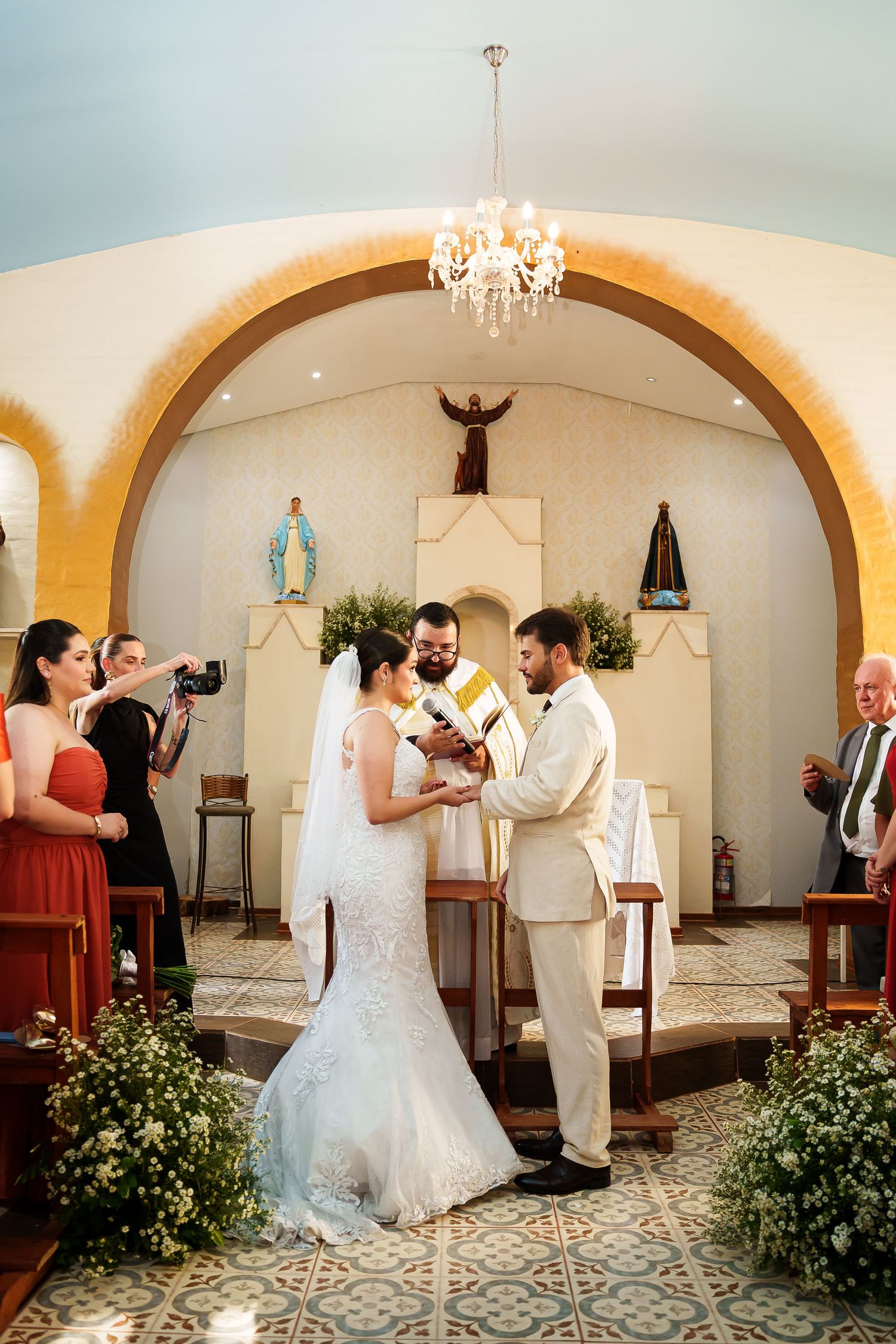 Bride and groom exchange vows at a Catholic church altar with a priest, chandelier, and religious statues in the background.