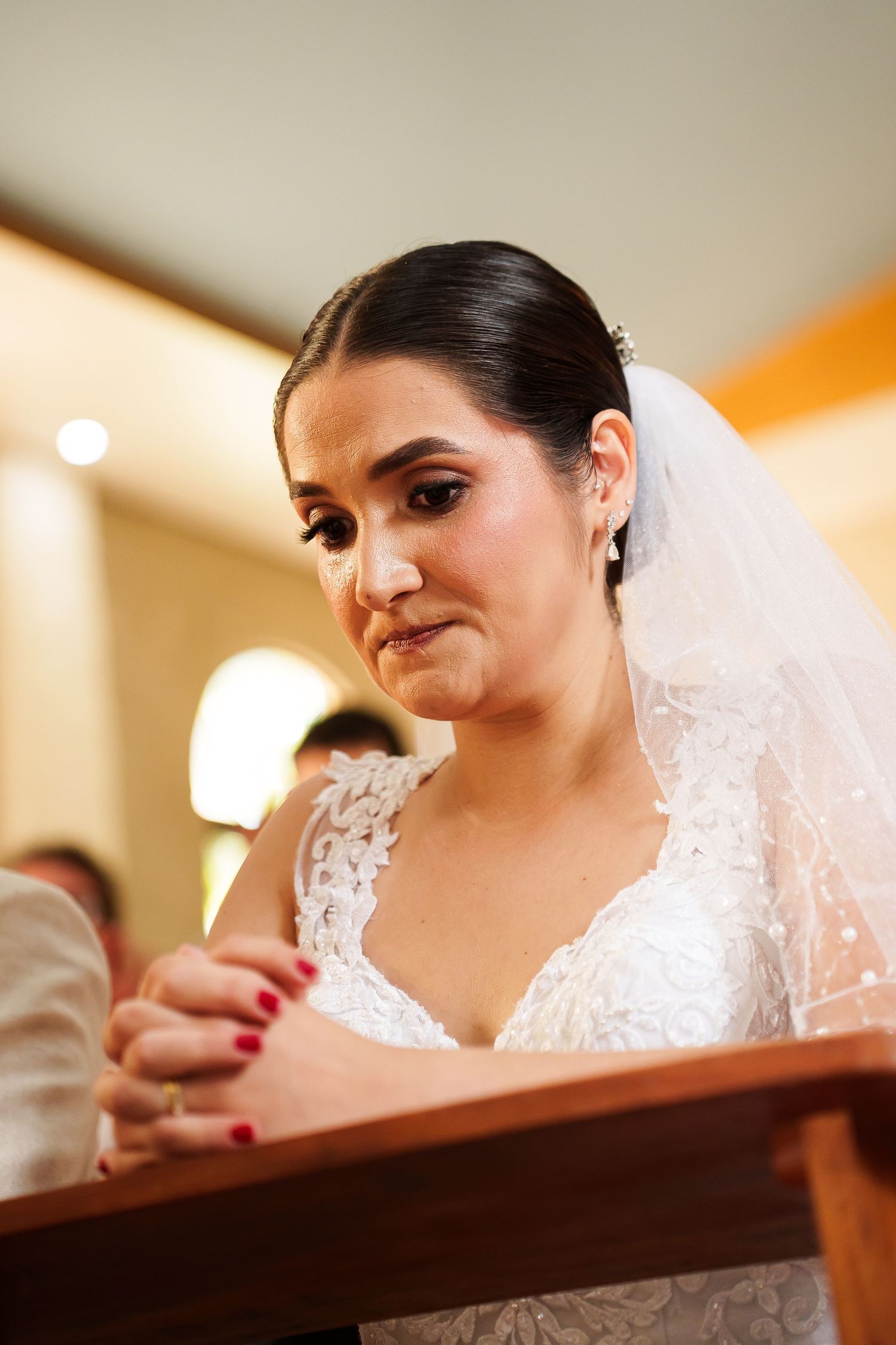 Emotional bride in white lace gown and veil praying at church altar during wedding ceremony.