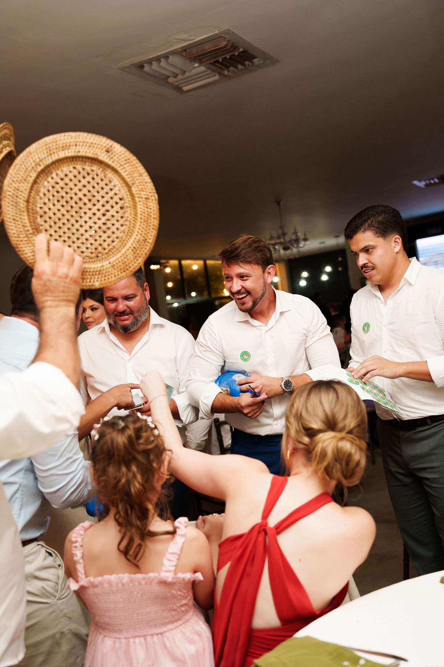 Group of people in white shirts celebrating at a wedding reception, laughing and exchanging gifts indoors.