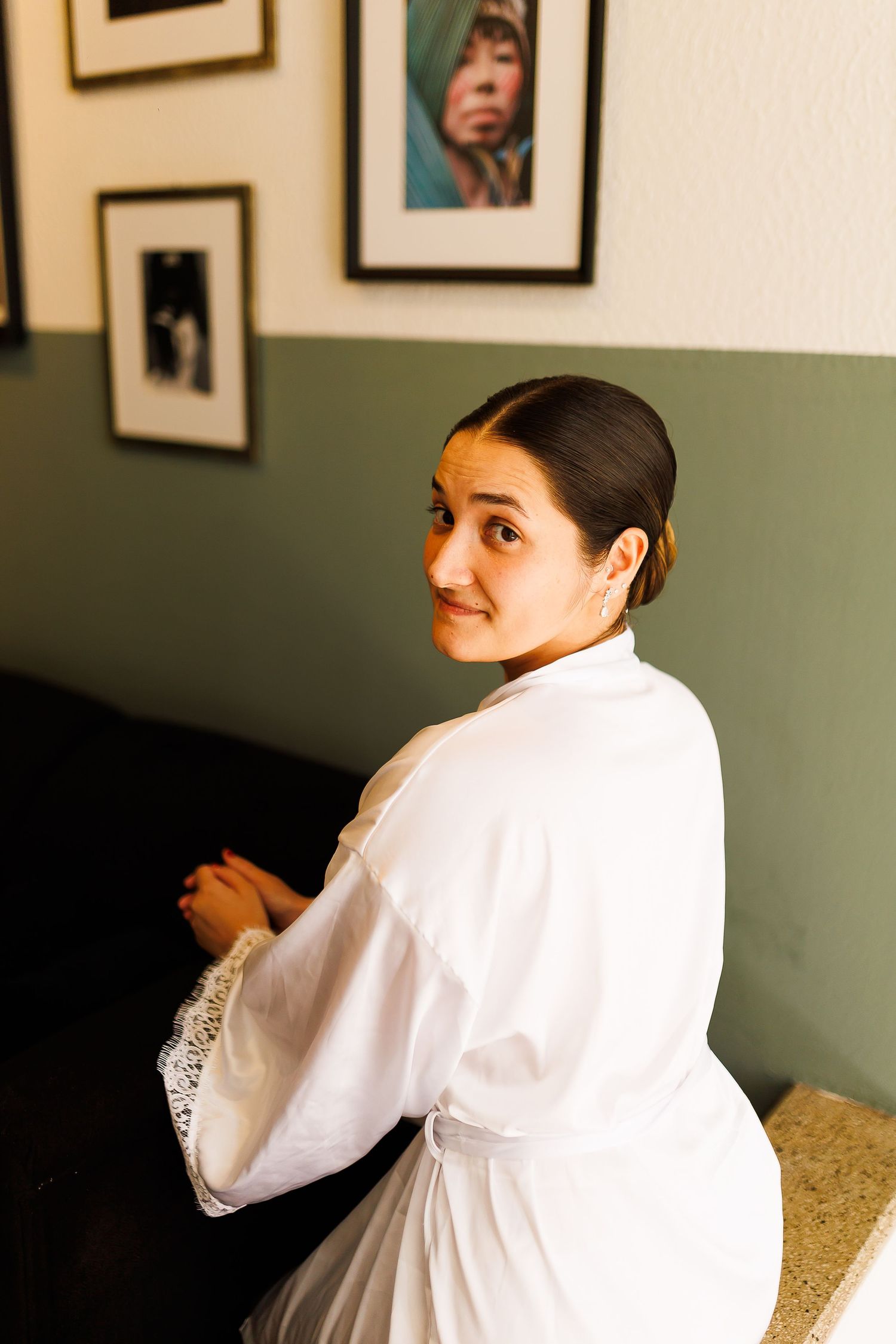 Bride in white robe glancing back over shoulder, elegant updo hairstyle, framed art on sage green wall.