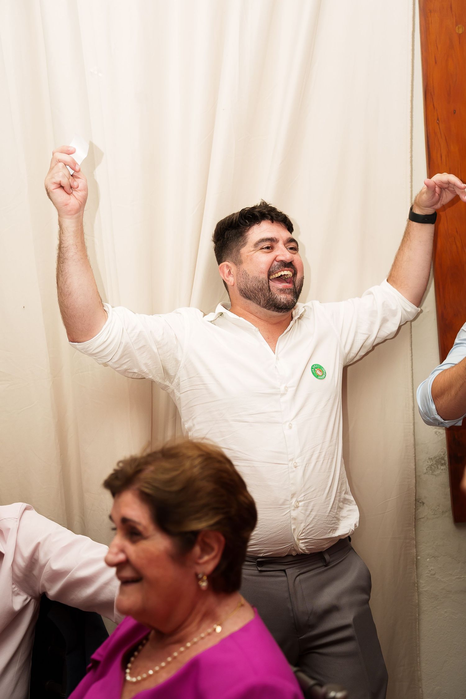 Excited man in white shirt raises arms joyfully at a social gathering, with a woman in pink blouse in the foreground.
