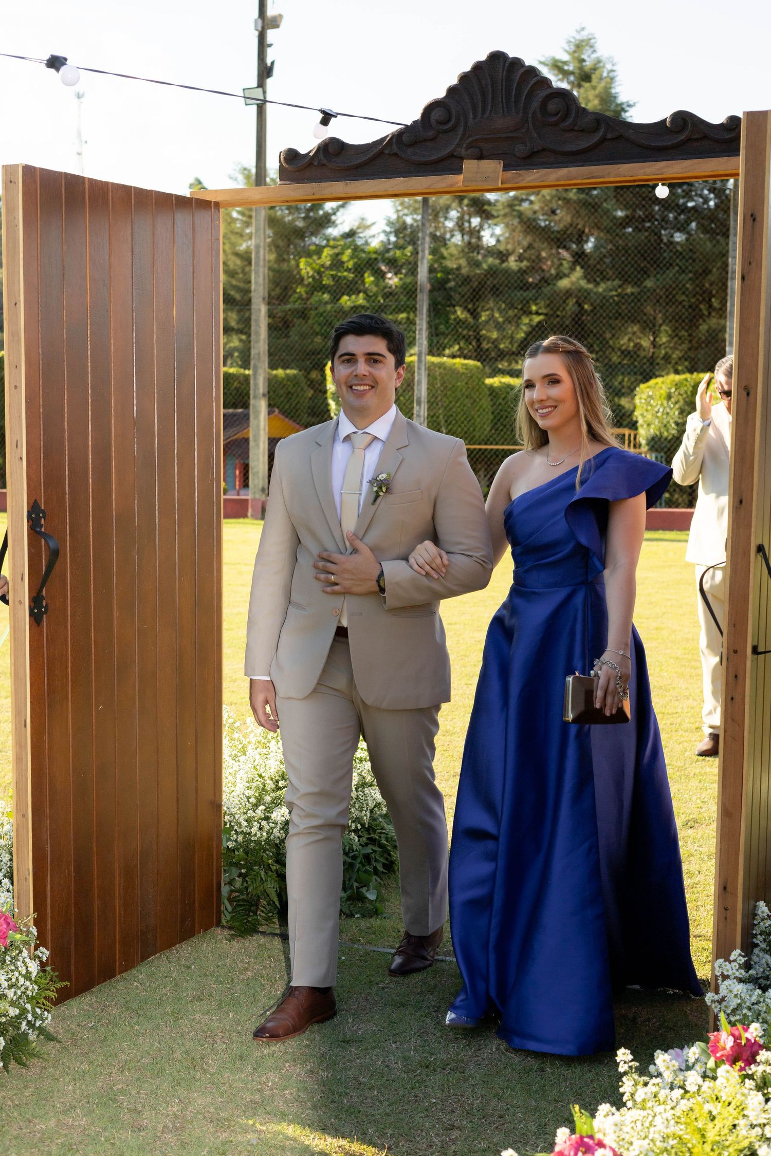 Elegant couple at outdoor wedding, man in beige suit and woman in royal blue one-shoulder gown walking through wooden arch.