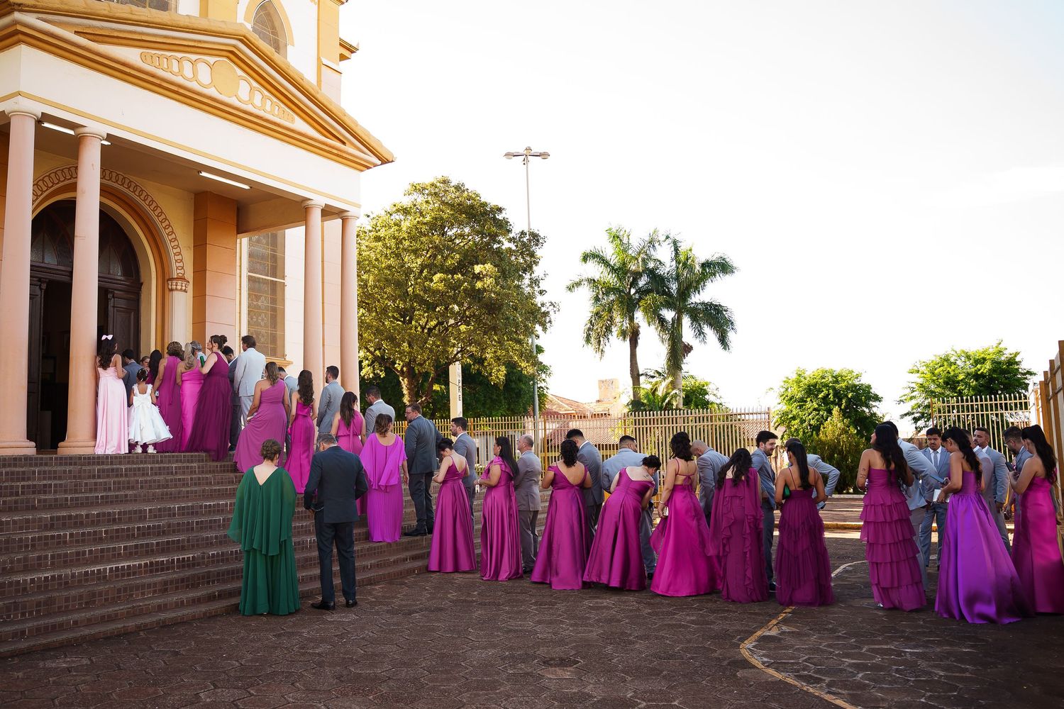 Wedding party in magenta and green gowns gathered outside a golden church with palm trees at sunset.