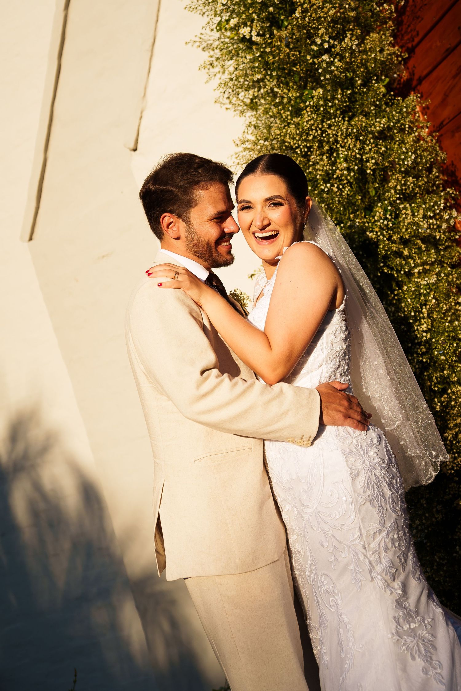 A joyful bride in a white lace gown and veil embraces her groom in a beige suit outdoors in golden sunlight.