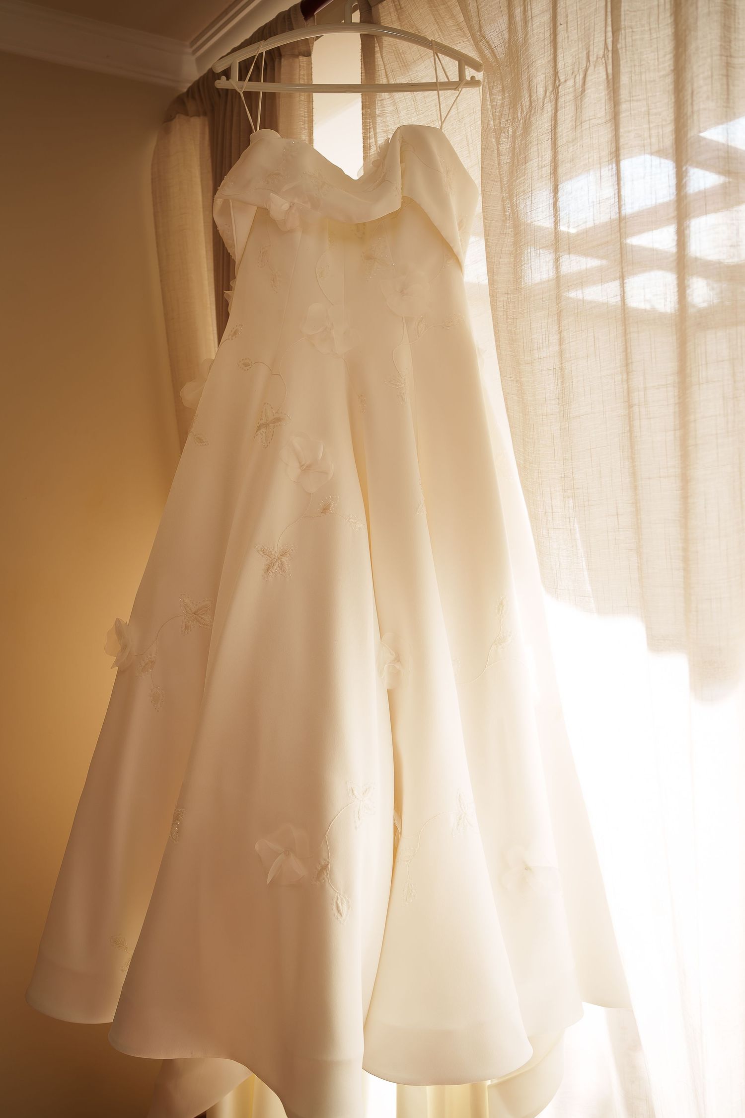 Elegant white strapless A-line wedding dress with floral appliques hanging on a hanger near a sunlit window.