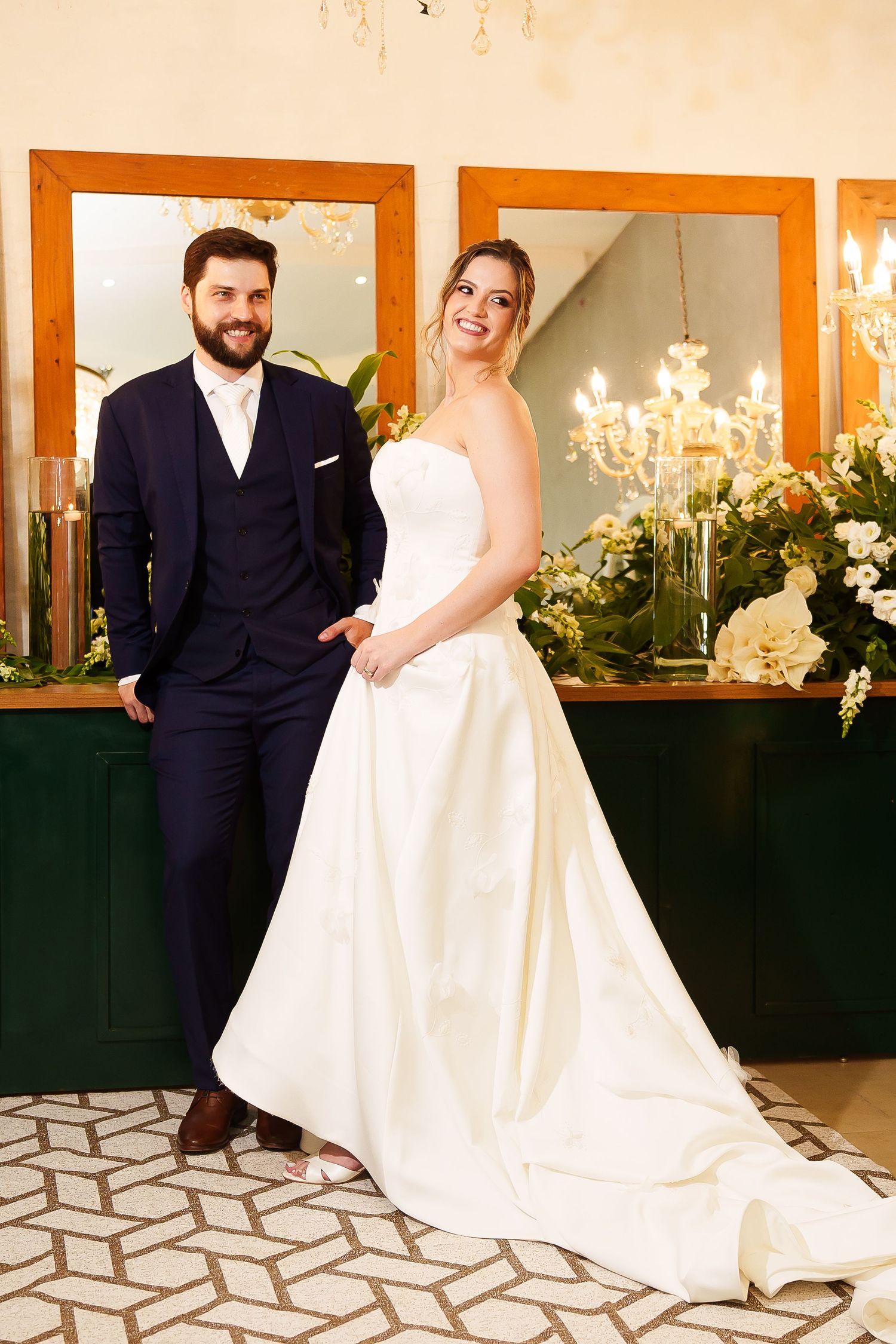 Elegant bride in white strapless gown and groom in navy suit posing together at a decorated wedding venue.