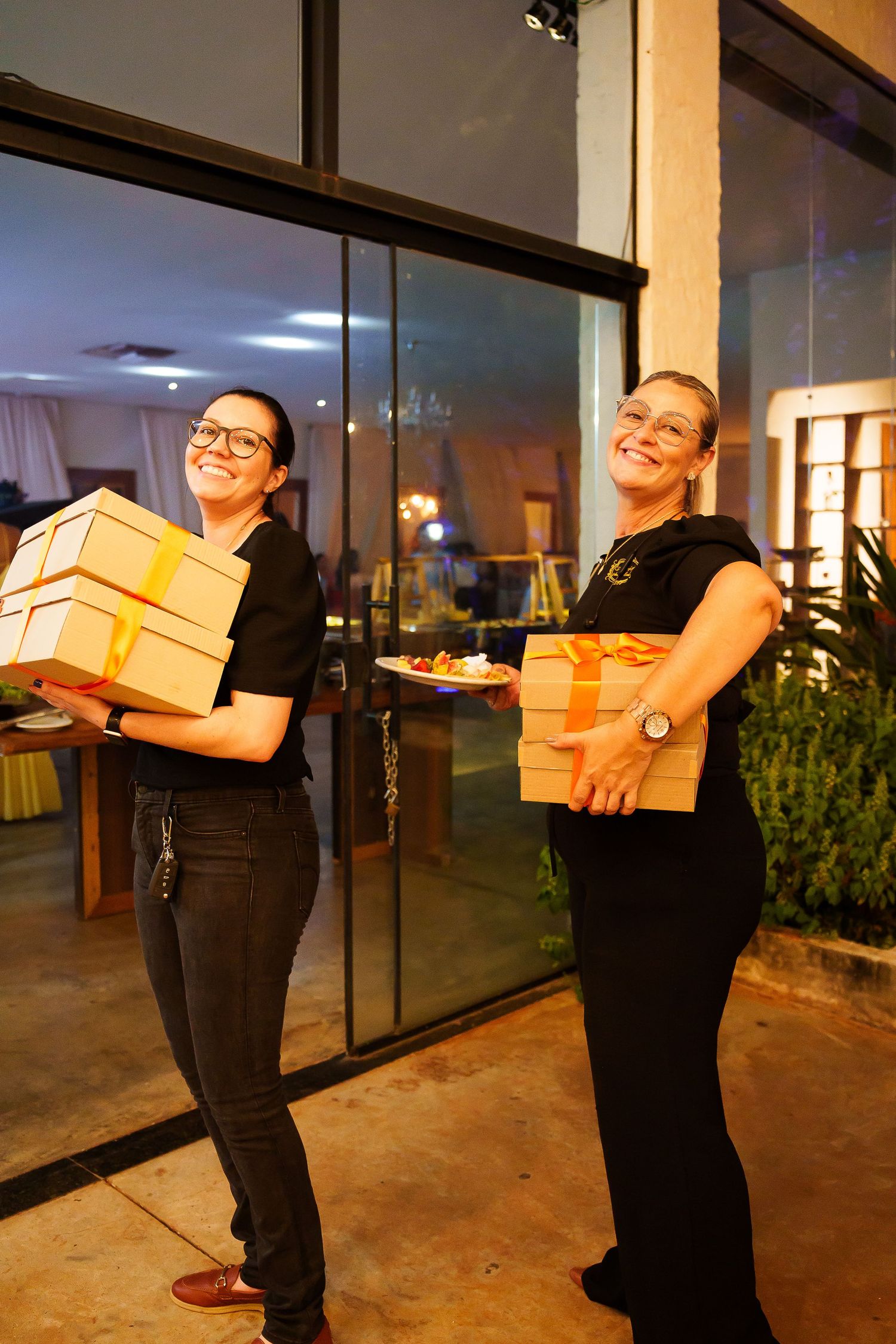 Two women in black outfits smiling while holding gold gift boxes outside an event venue.