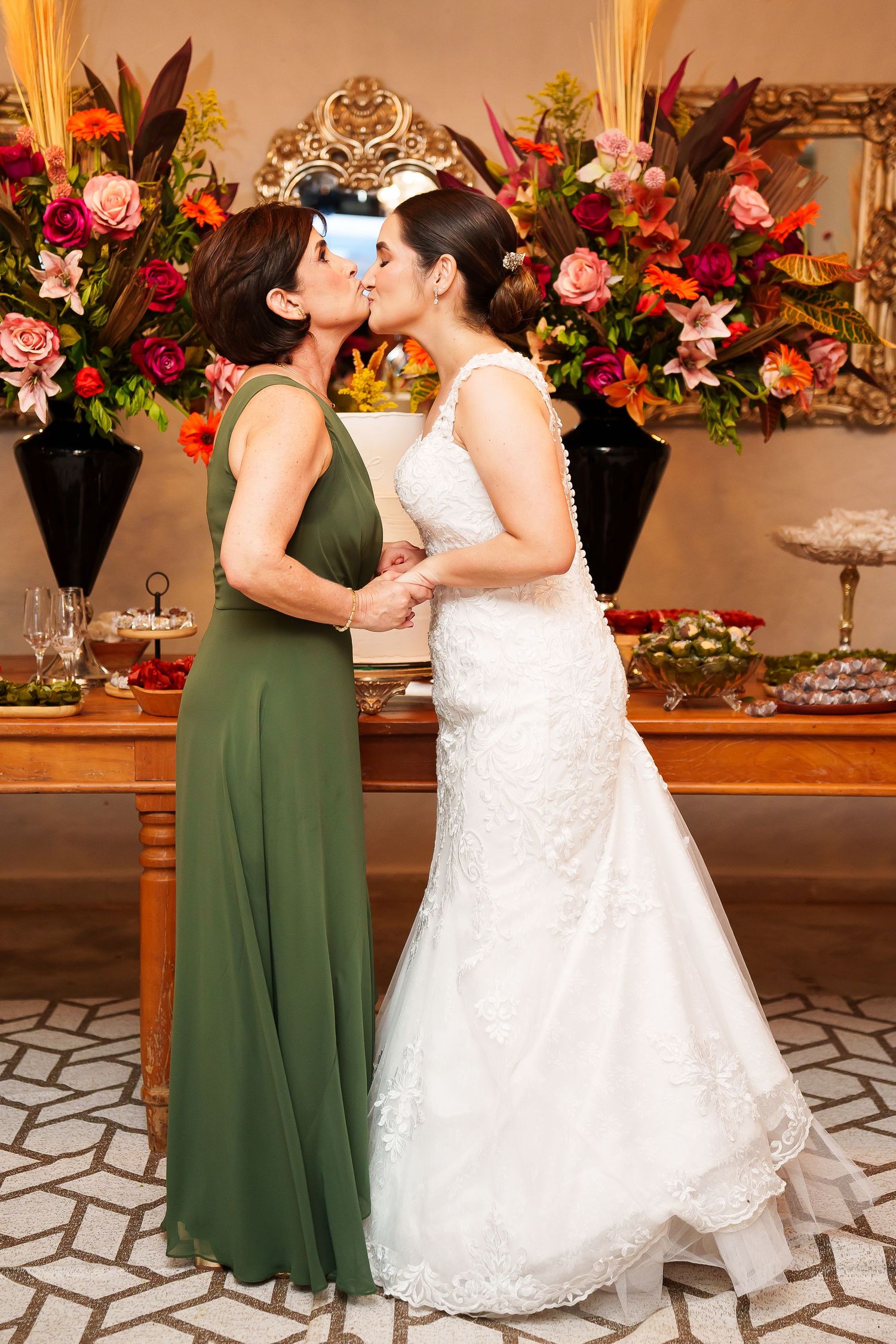 Bride in white lace gown shares a kiss with woman in green dress at elegant floral wedding reception.