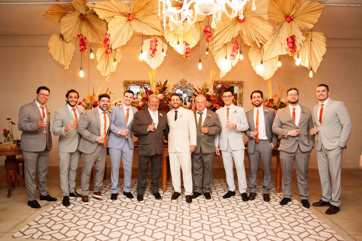 Groom in white suit poses with groomsmen in gray suits and orange ties at elegant wedding venue.