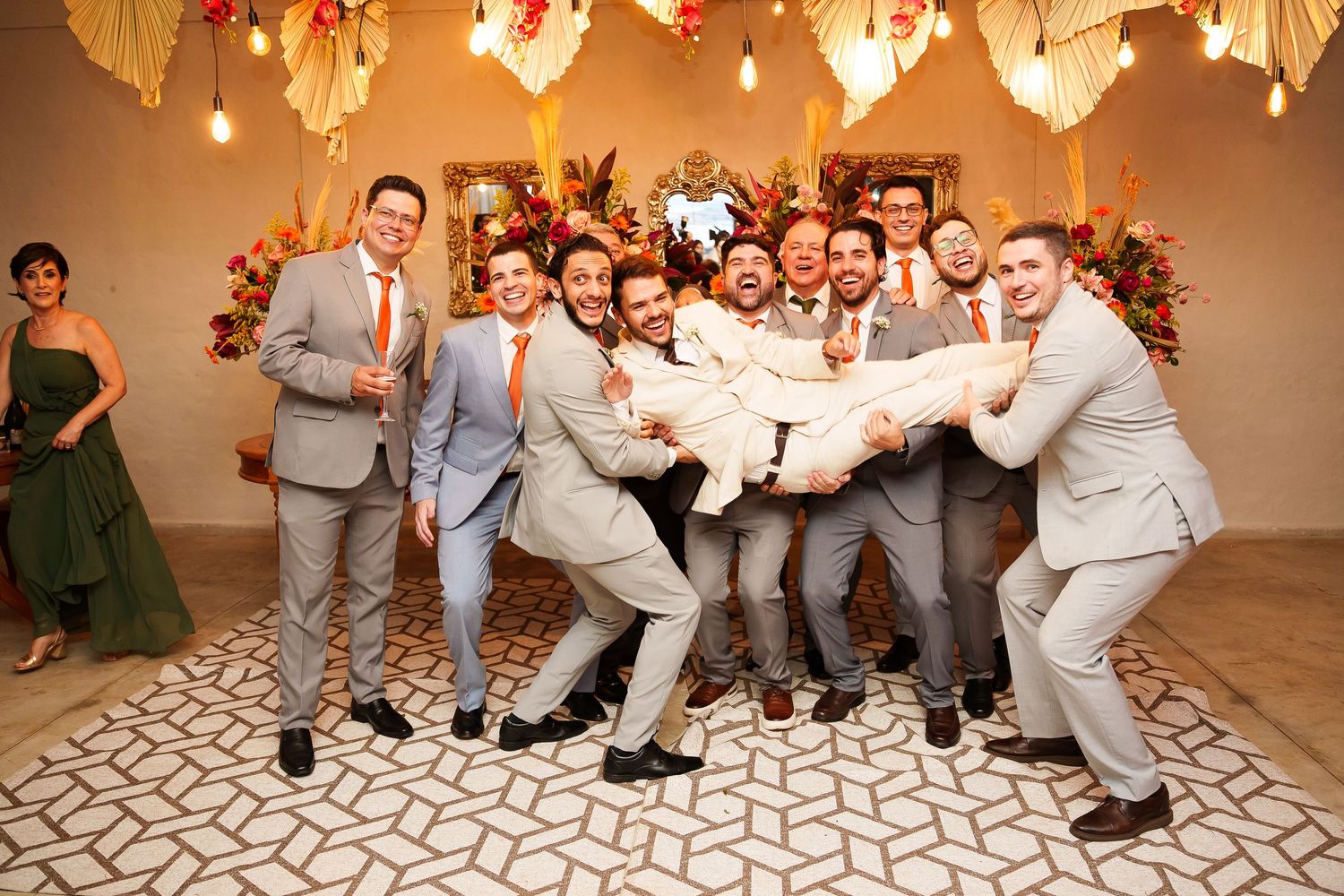 Groomsmen in gray suits lift the groom at a festive wedding reception with colorful floral decor.