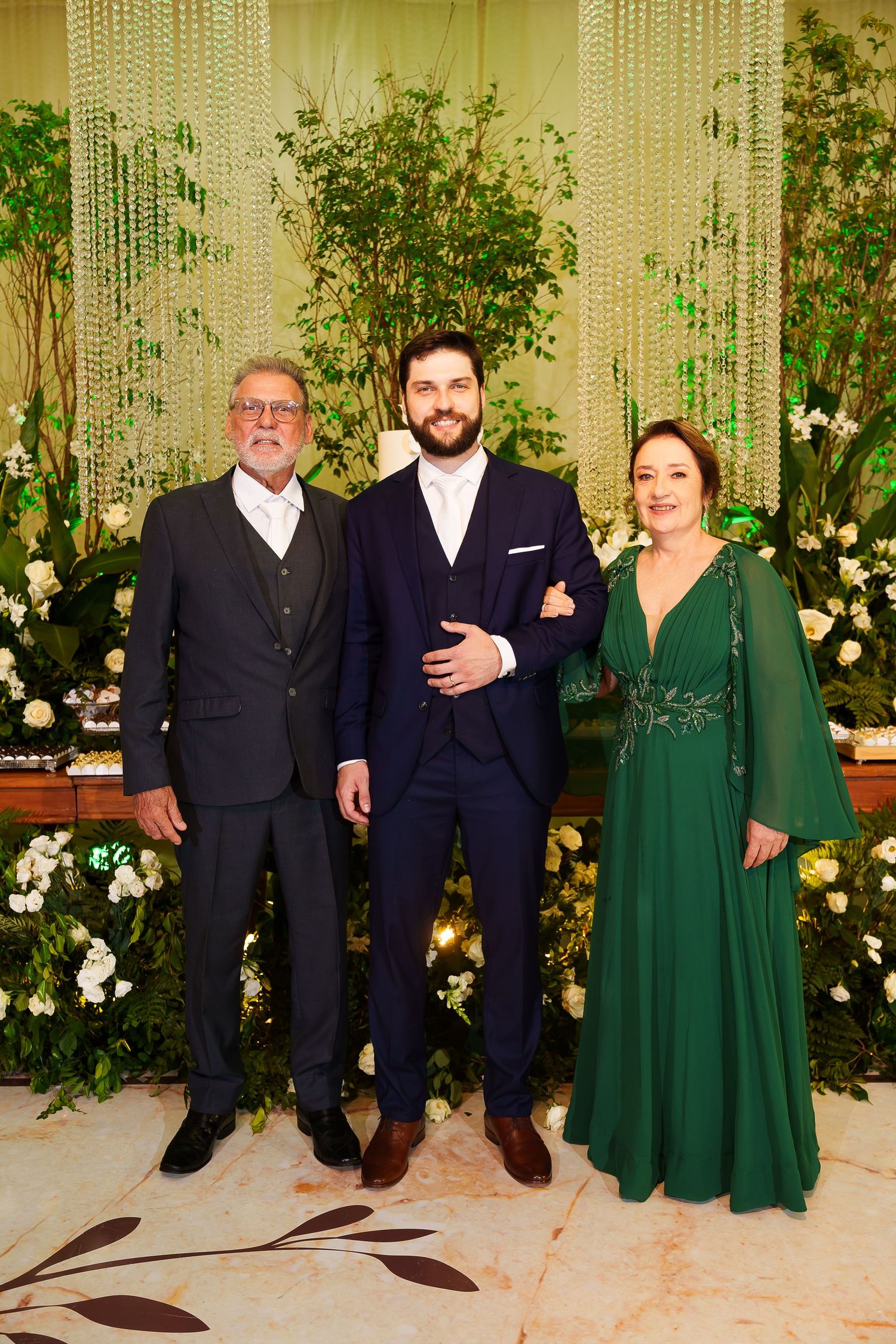 Three elegantly dressed people pose together at a formal event, with the woman wearing a stunning emerald green gown.