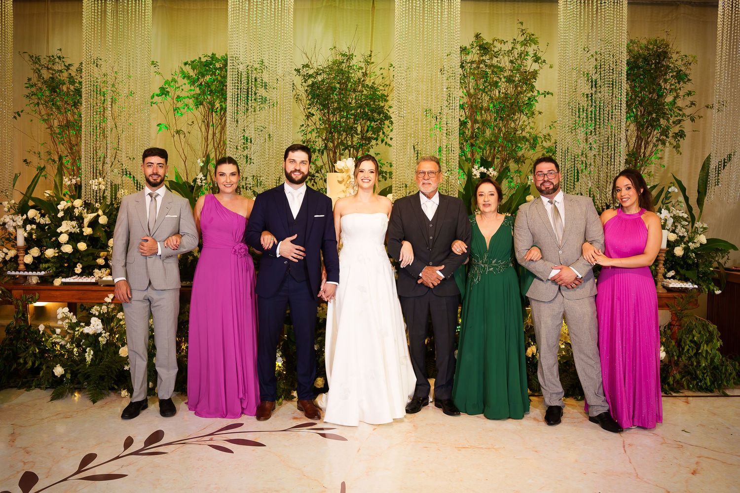 Wedding party poses together in elegant venue with lush greenery, bride in white gown flanked by guests in purple, green, and gray.