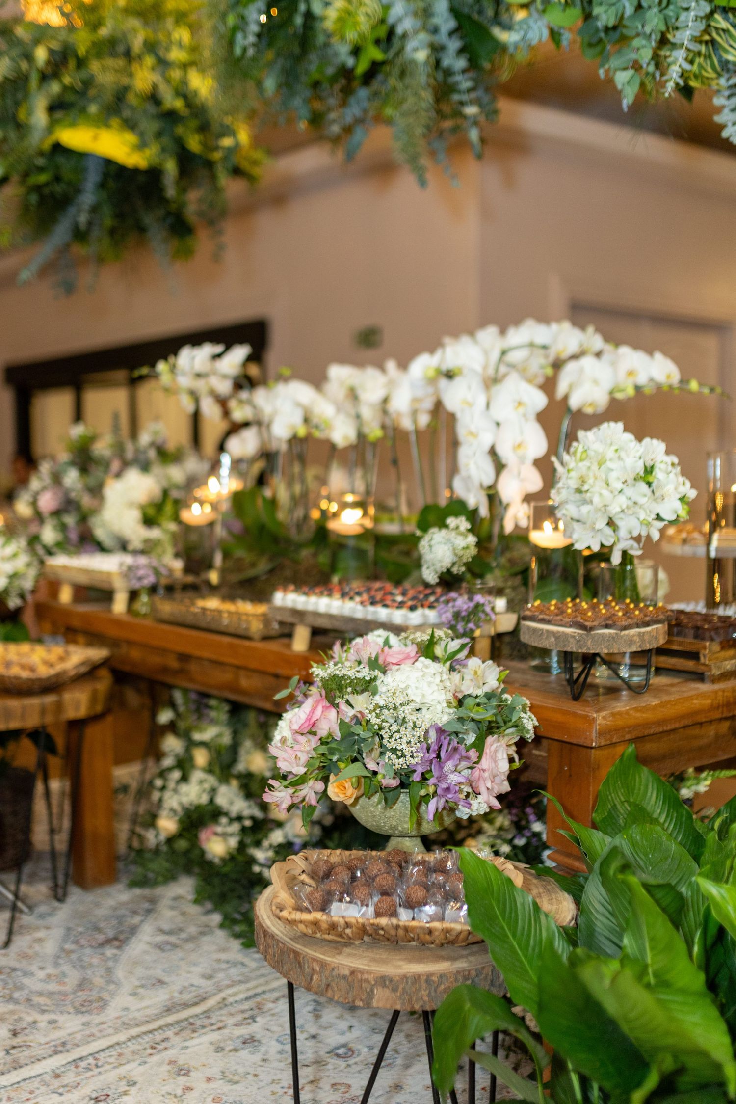 Elegant wedding dessert table with white orchids, candles, and lush greenery arrangements on rustic wooden tables.