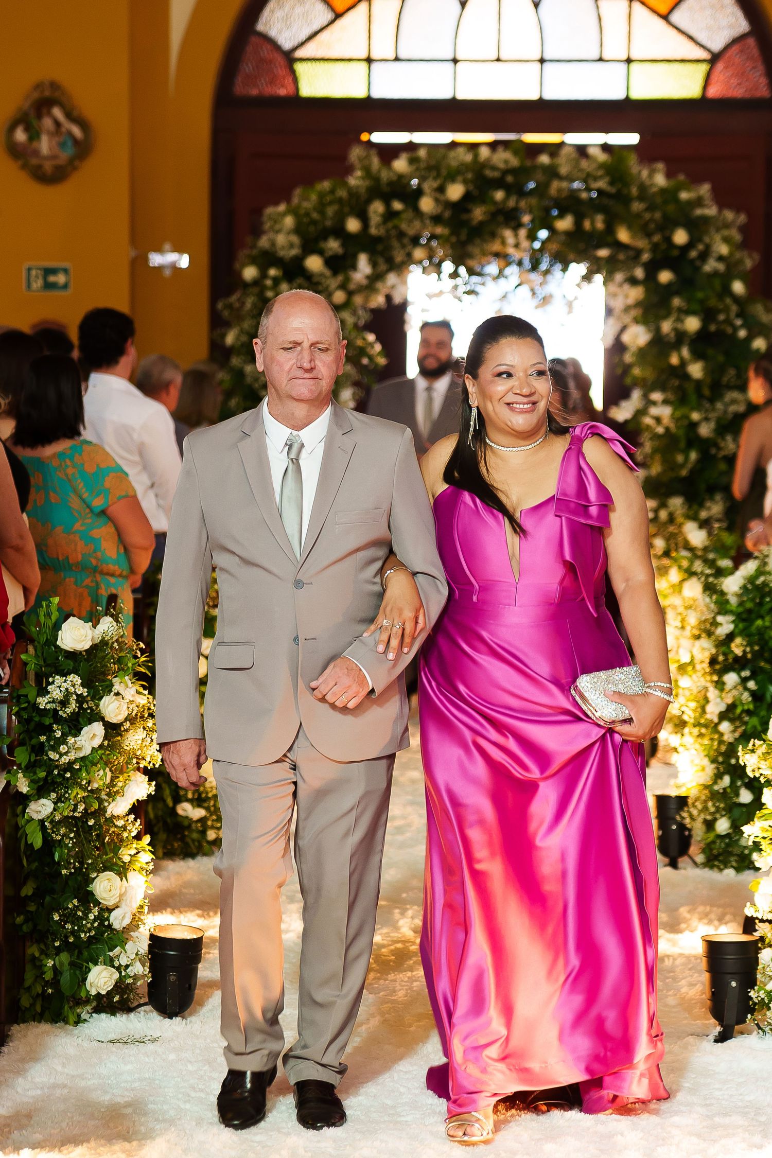 Elegant couple walking down a floral wedding aisle, man in tan suit and woman in vibrant pink gown.