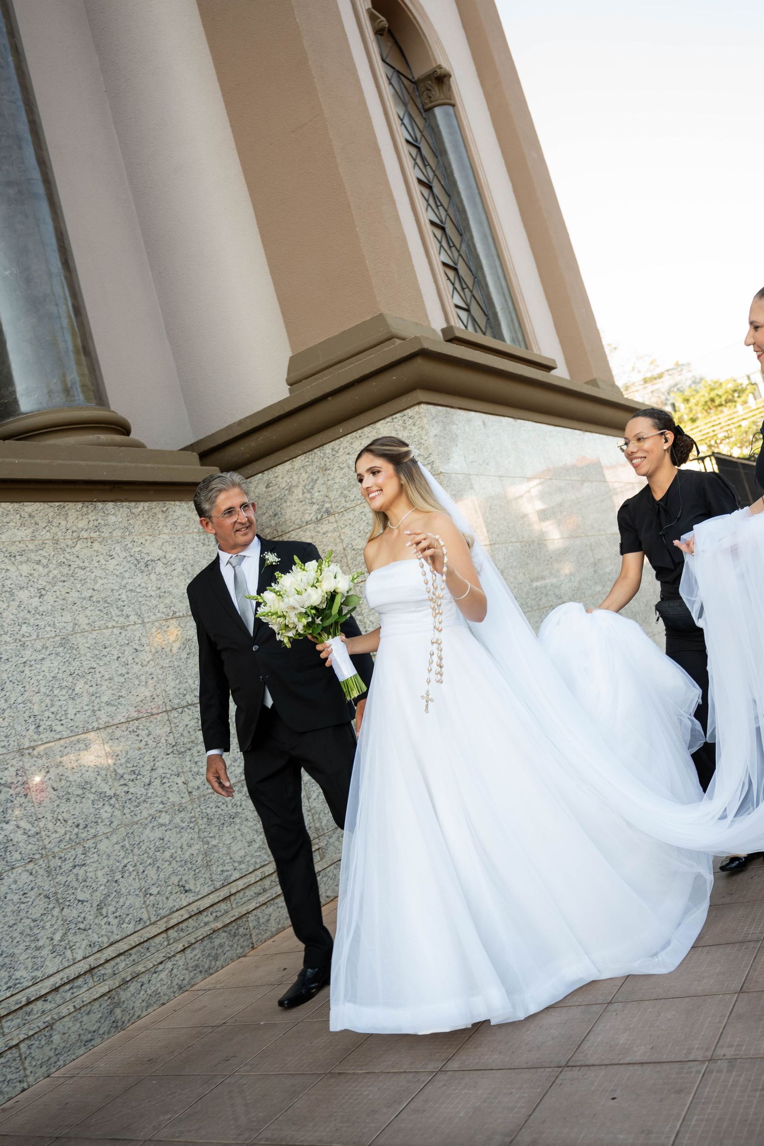 Bride in white gown holding bouquet walks with groom in black tux outside church, attendants helping with veil.