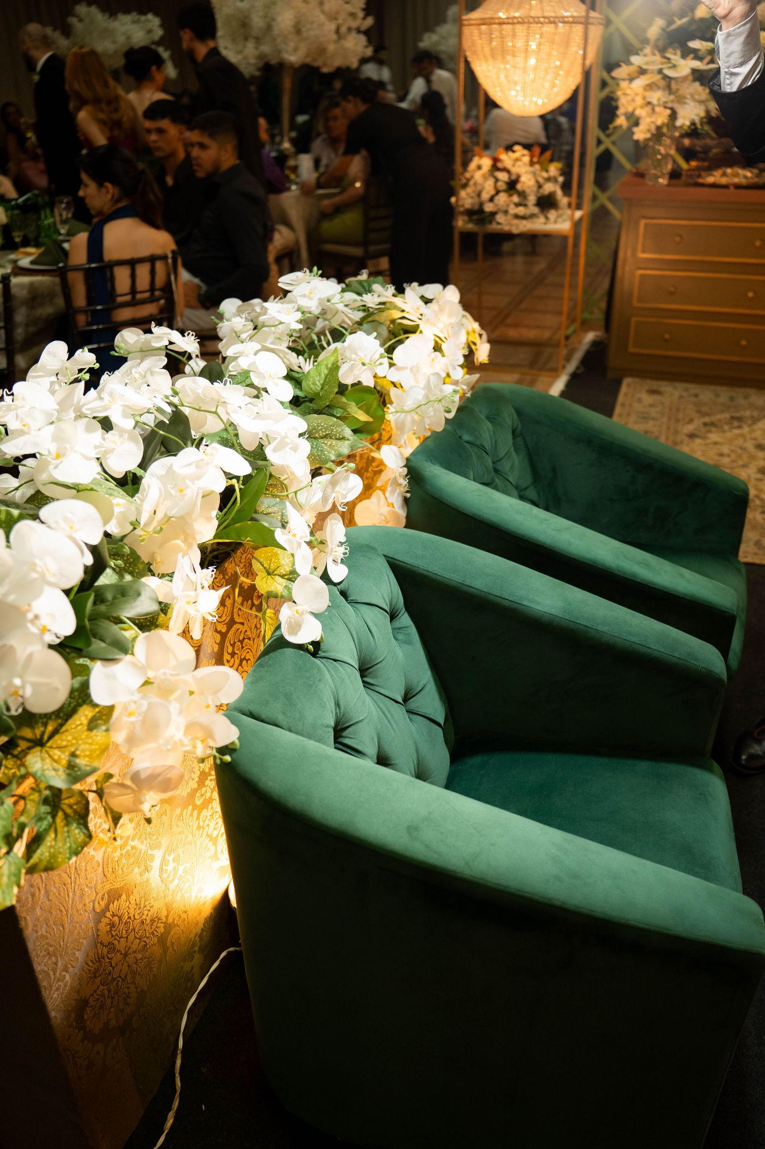 Elegant green velvet tufted chairs beside white floral arrangement at a formal event venue with warm lighting.