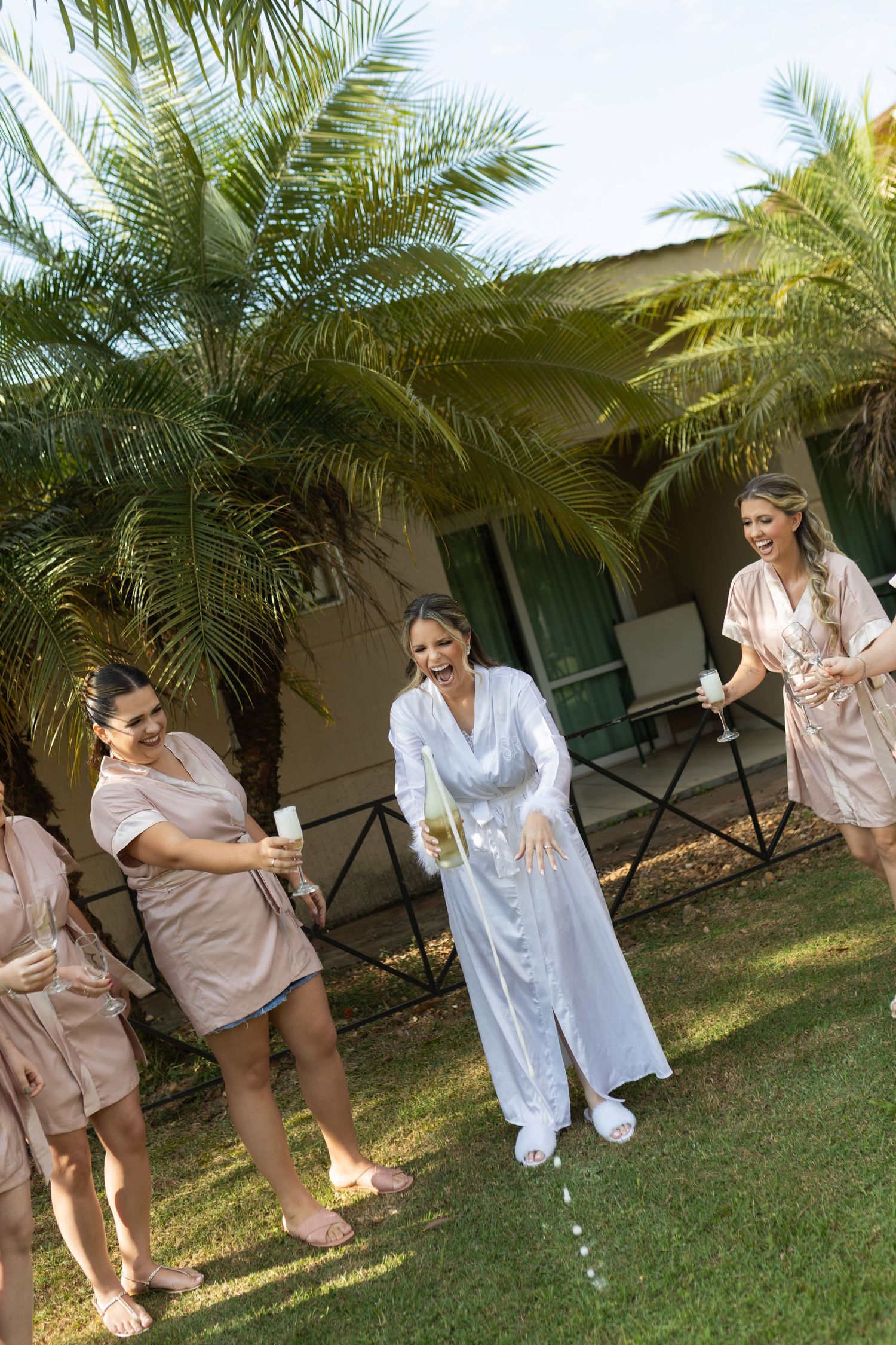 Bridesmaids in pink robes popping champagne with bride in white robe outdoors near palm trees.