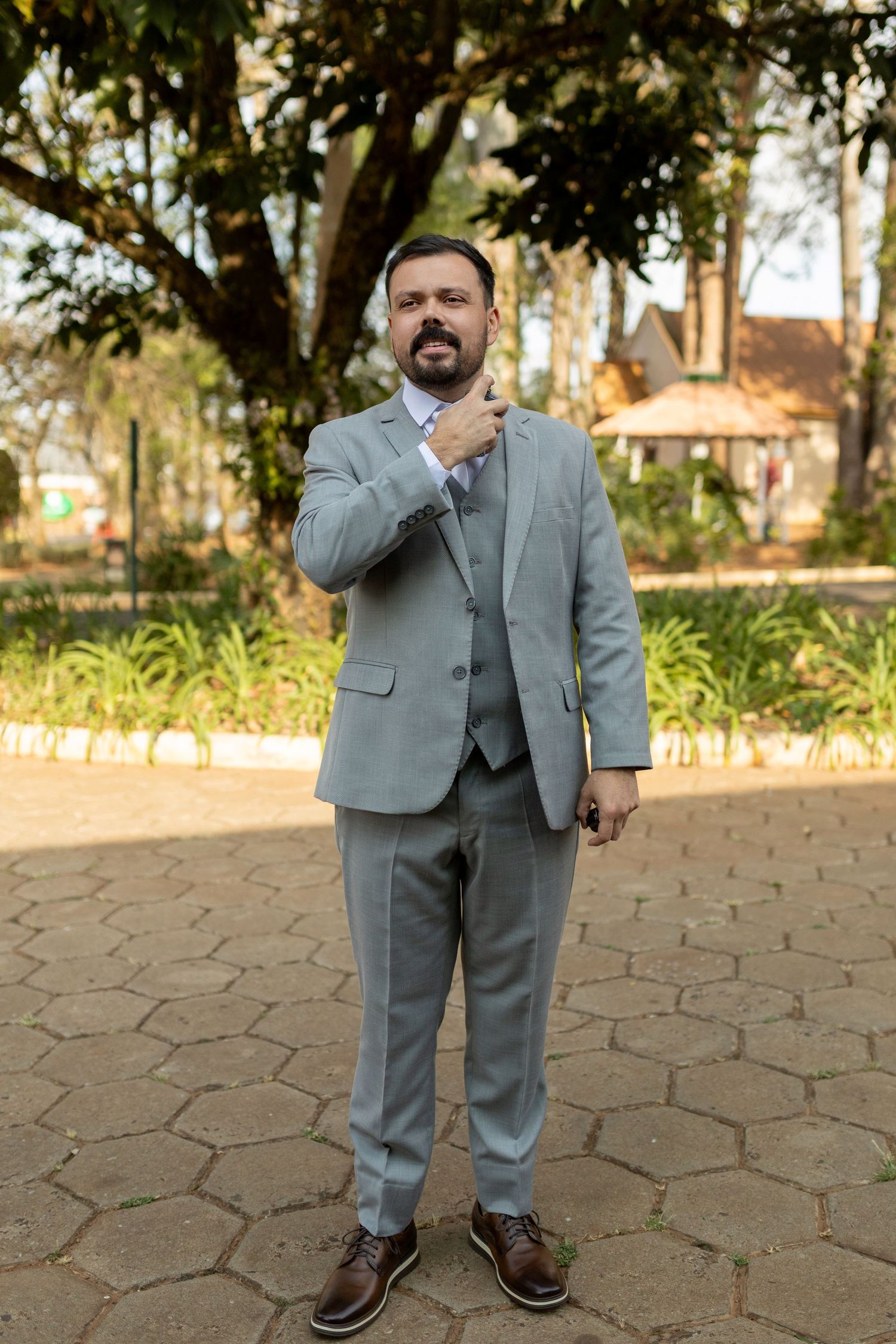 Man in gray three-piece suit adjusting tie in outdoor park setting with trees and brick pathway.