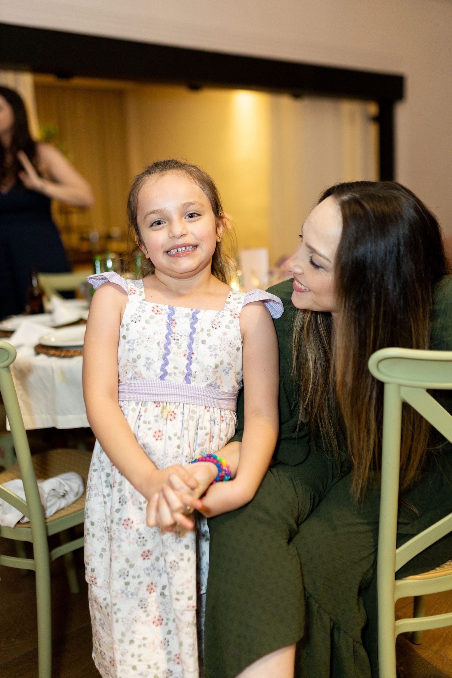 A young girl in a floral dress smiles at the camera while a woman in green looks at her lovingly at a dinner event.