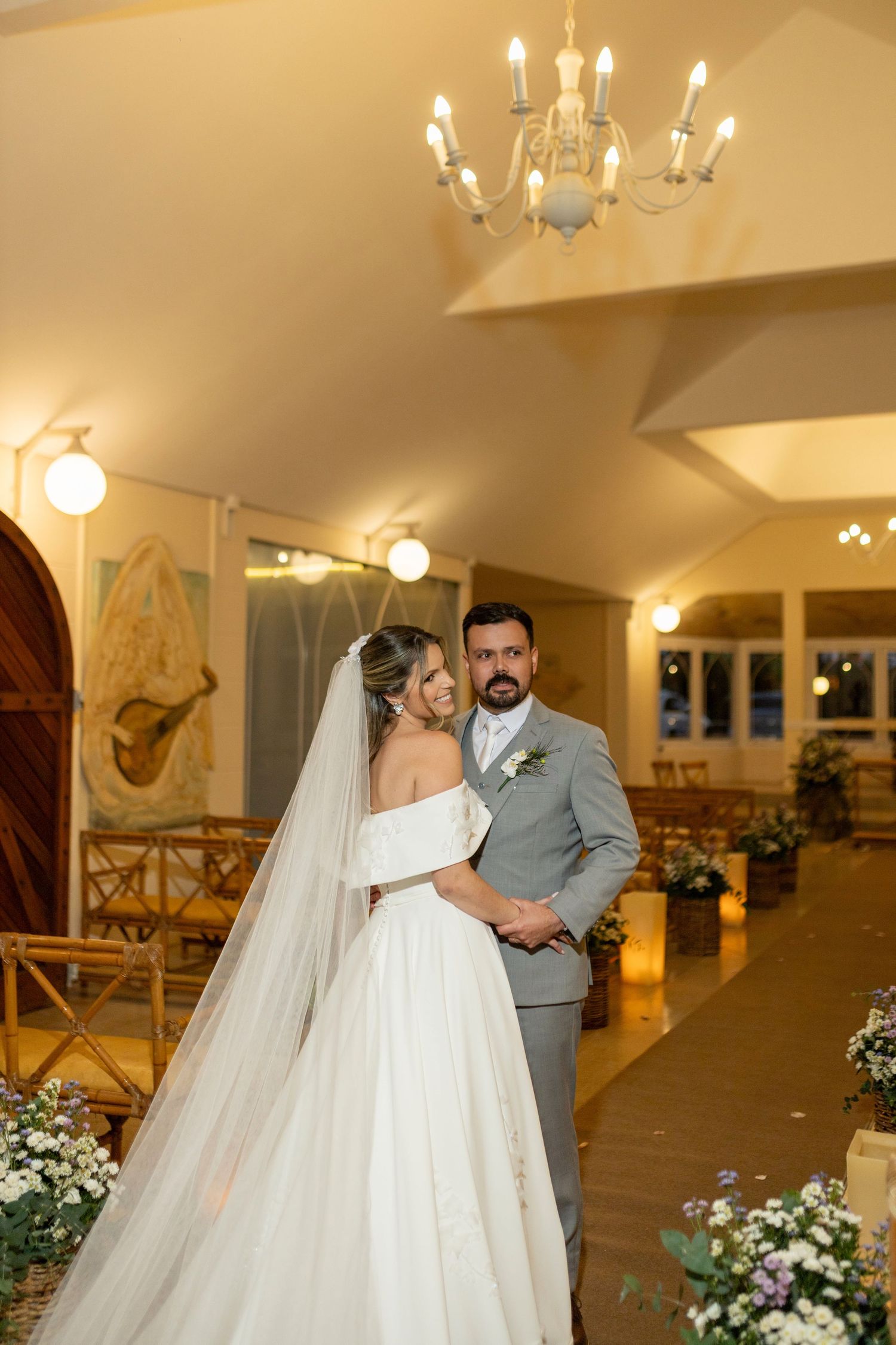 Bride in off-shoulder gown with veil embraces groom in gray suit inside warmly lit wedding chapel.