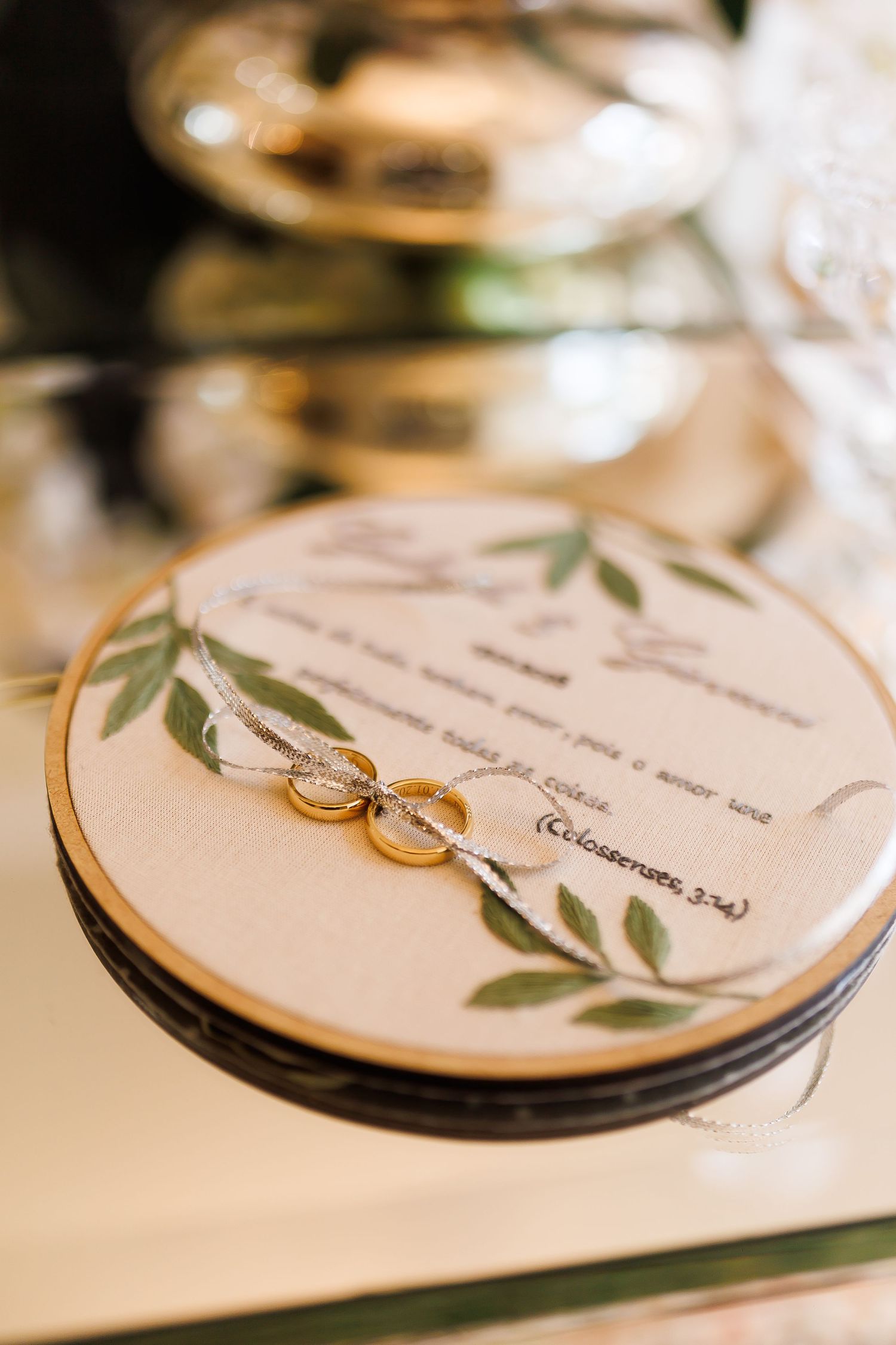 Two gold wedding rings resting on an embroidered hoop with green leaves and a Bible verse Colossians 3:14.