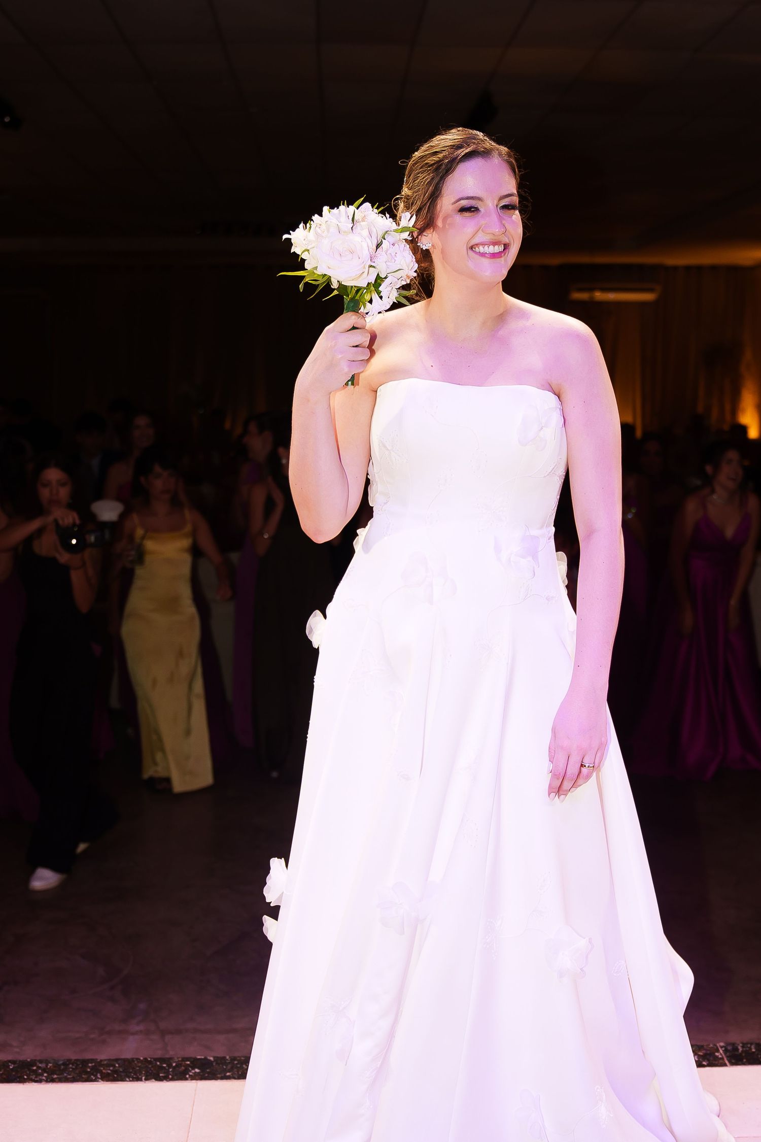 Smiling bride in strapless white gown holding bouquet of white flowers on dance floor at wedding reception.
