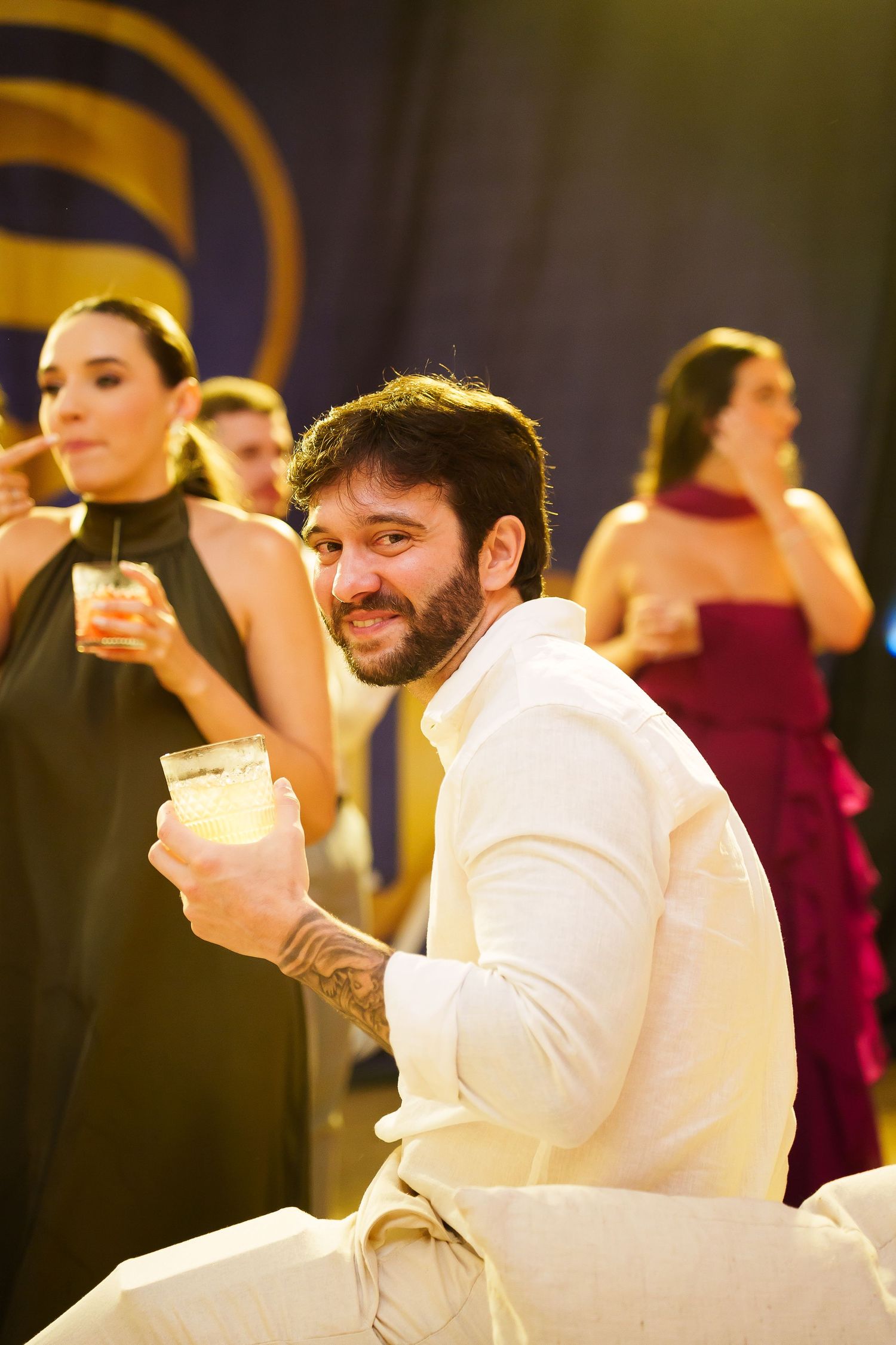 Smiling bearded man in white suit holding a cocktail glass at an elegant evening event.