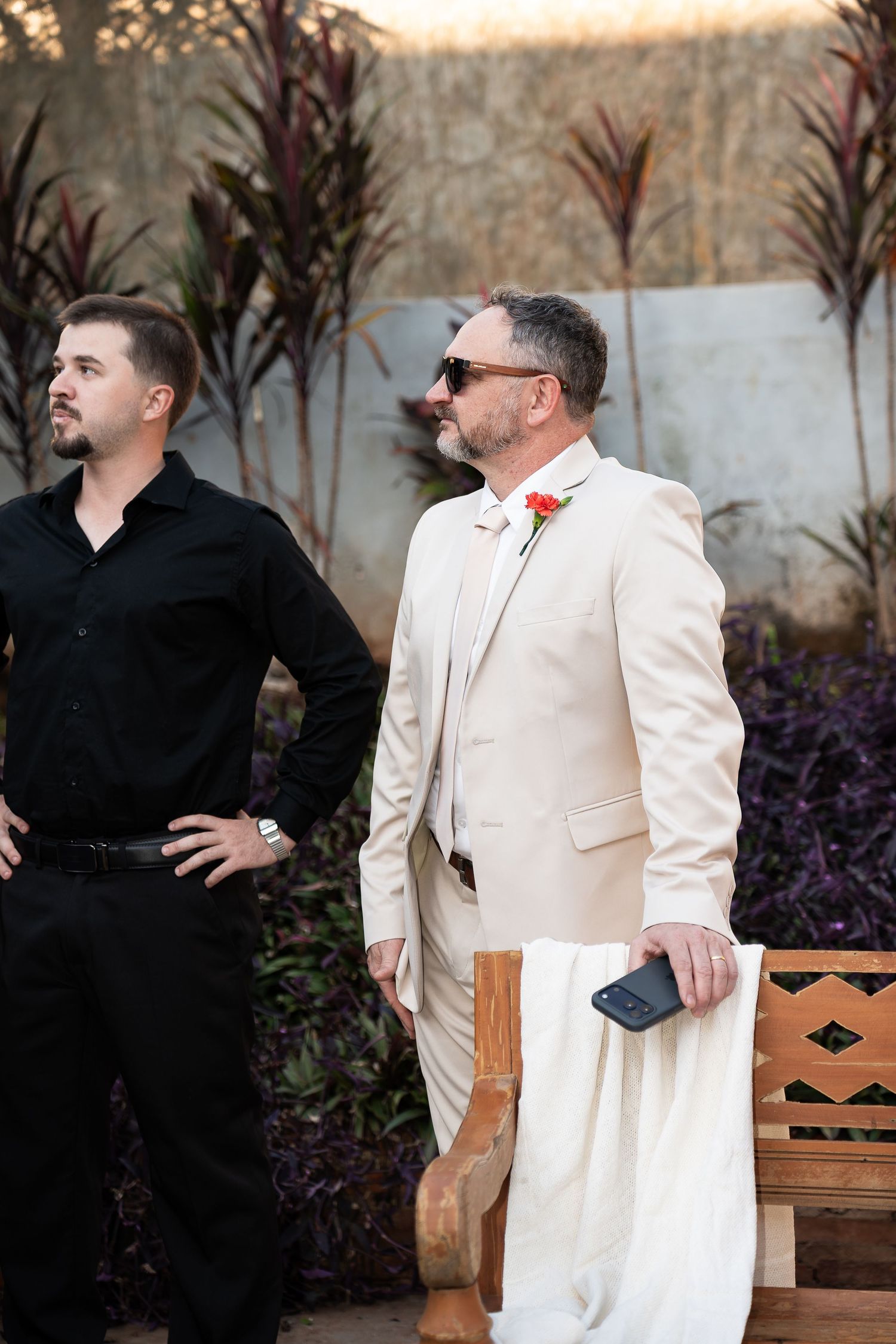 Groom in cream suit with red boutonniere stands beside man in black at outdoor wedding ceremony.